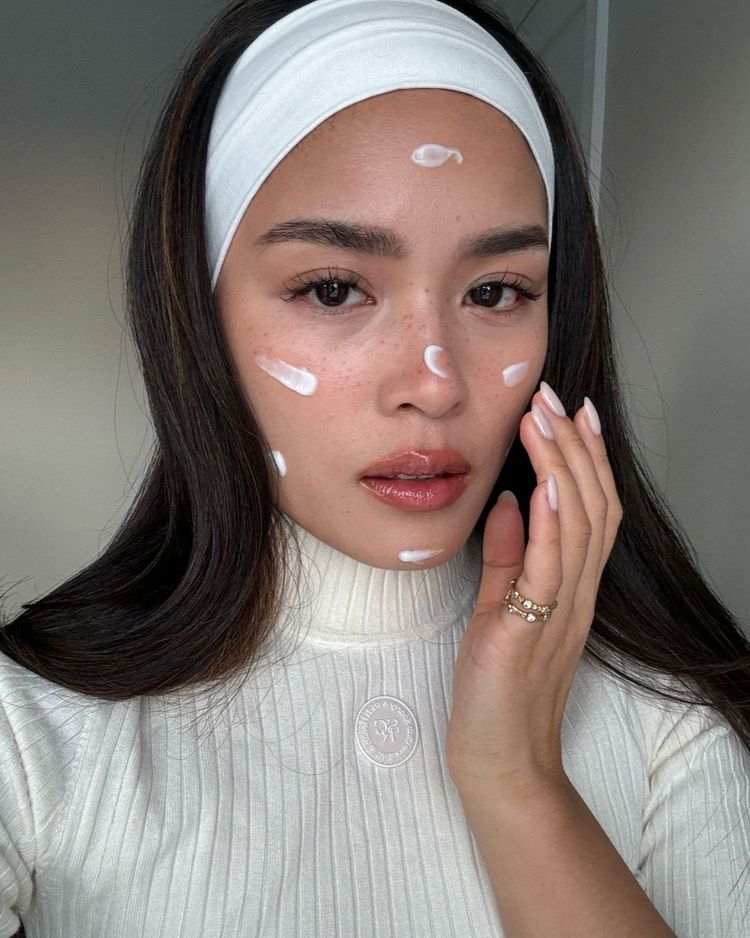 A portrait of Tsutsumi Hoang applying moisturiser to her face, her hair pushed back by a white hairband