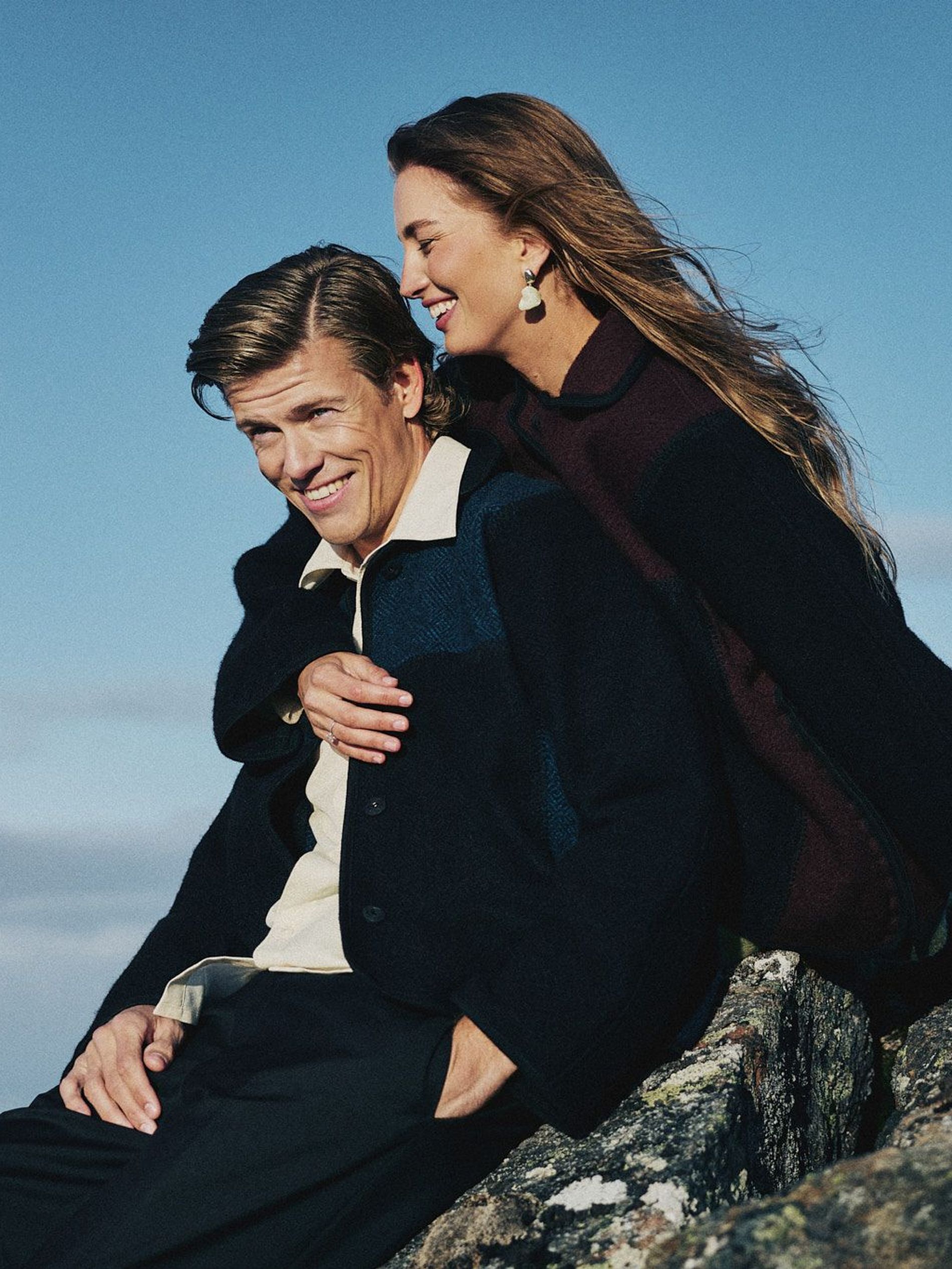 Klæbo and Døsvik sit on a rock. He wears black trousers and jacket with denim details with cream shirt. She wears all black