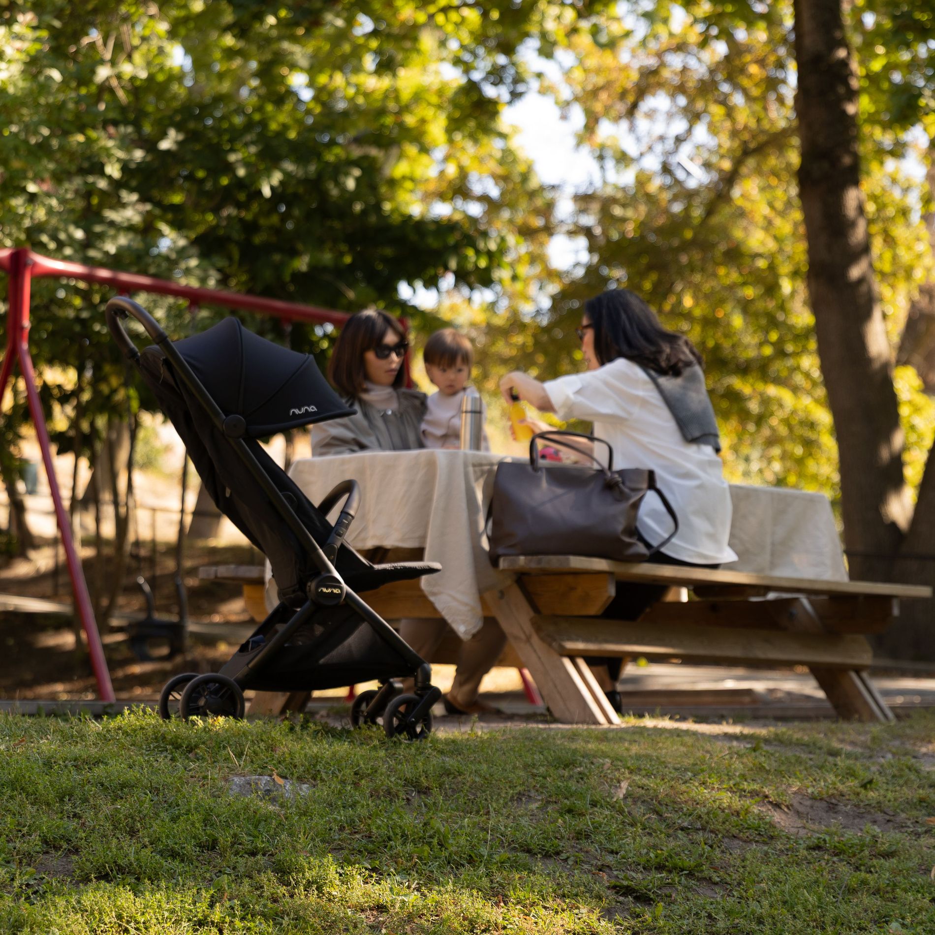 A grandmother, mother and child sit in the aprk having a picnic at a table, with the Nuna VIAA cabn nearby