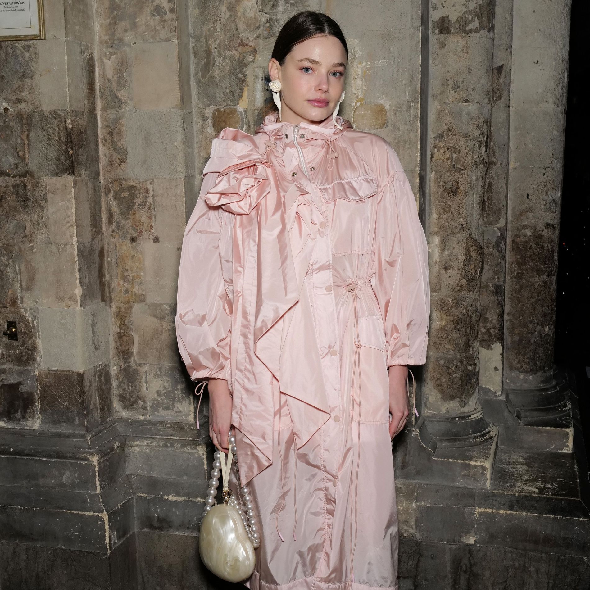 Kristine Froseth wears a pink raincoat with a bow-detailed shoulder, a pearl handbag and embellished crocs to the Simone Rocha AW24 show