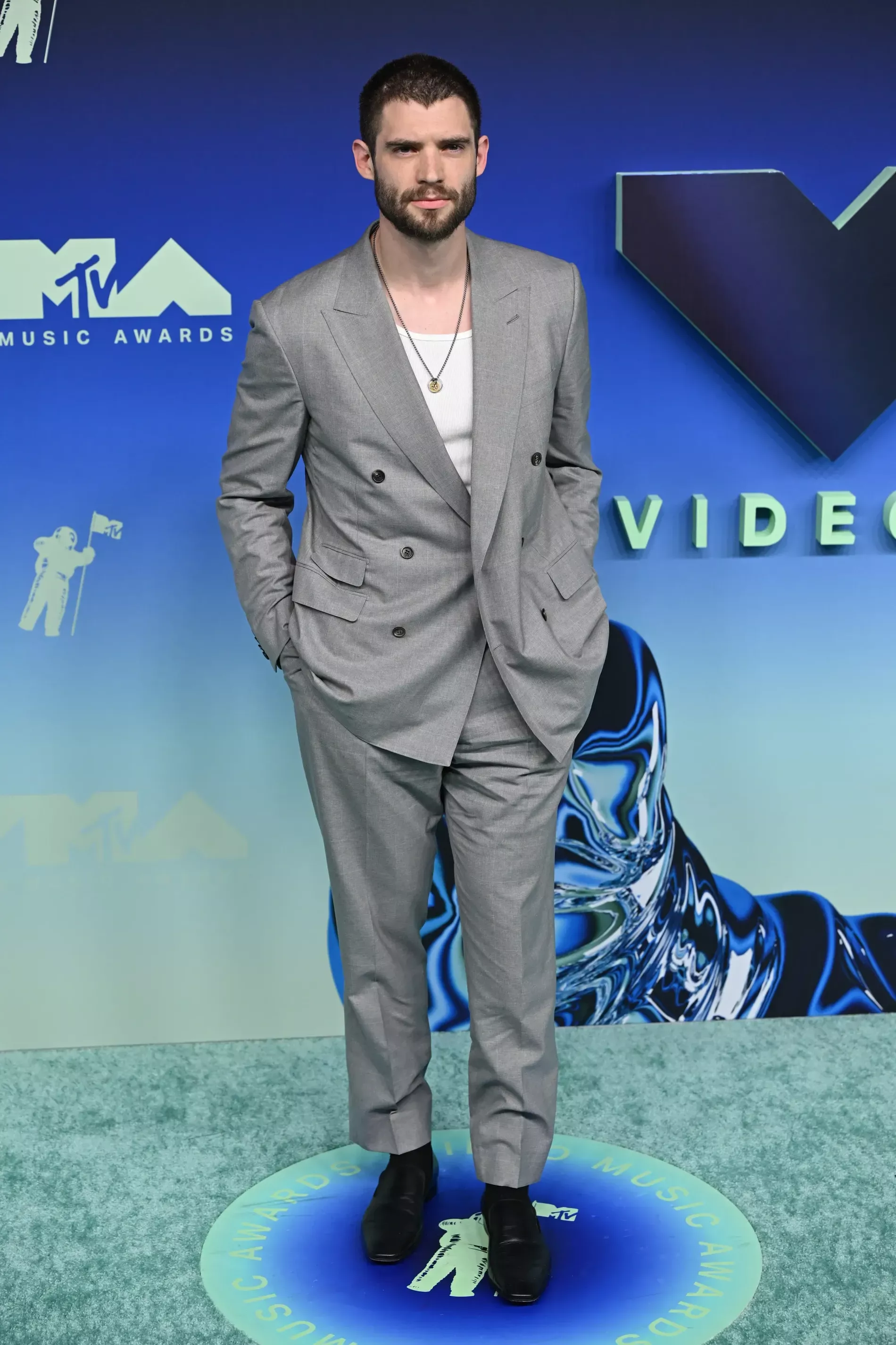 The image shows actor David Corenswet arriving at the 2026 MTV Video Music Awards at the Peacock Theater in Los Angeles, He wore a relaxed grey double-breasted suit layered over a white t-shirt, complemented by a pendant necklace and black dress shoes