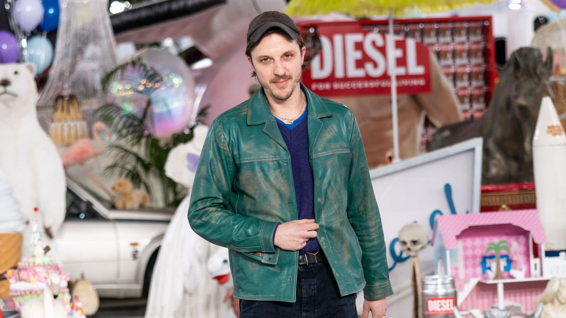 Glenn Martens wearing a green patrol leather jacket and dark blue jeans with brown shoes matching with a brown cap, he has alot of junk behind him from one of Diesels runway shows