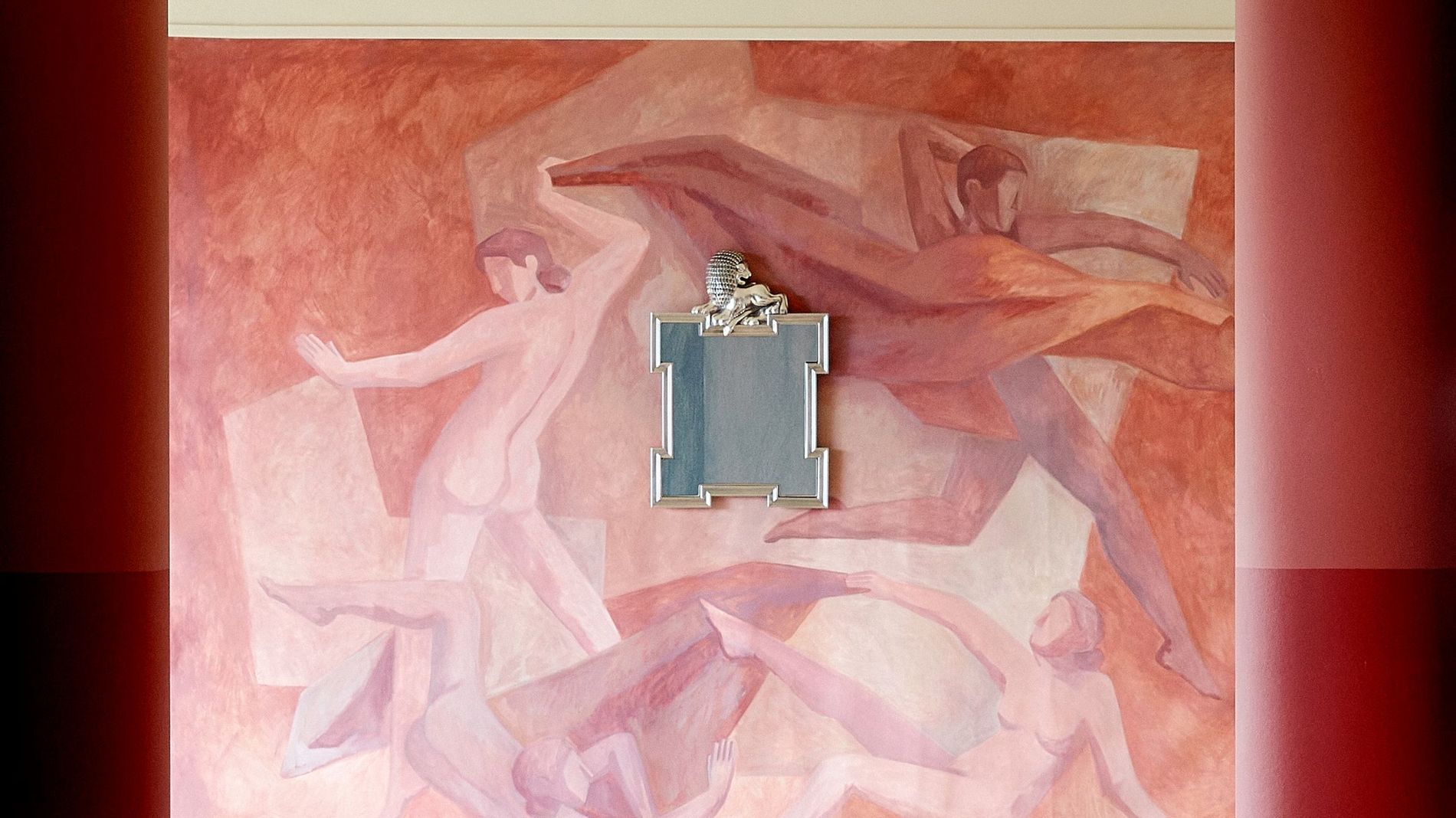 A red terracotta wall painted with naked people dancing and a silver mirror with a lion on the frame from Svenskt tenn Fall' 26