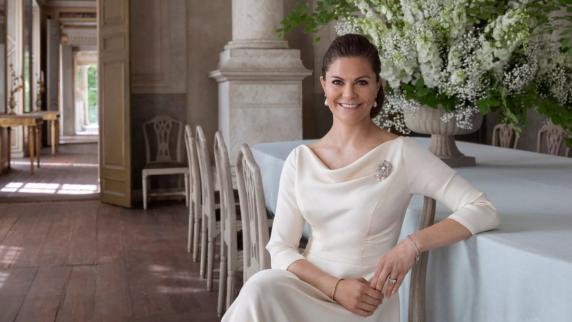 Crown Princess Victoria of Sweden poses sitting in a white gown with a draped neckline in a castle environment 