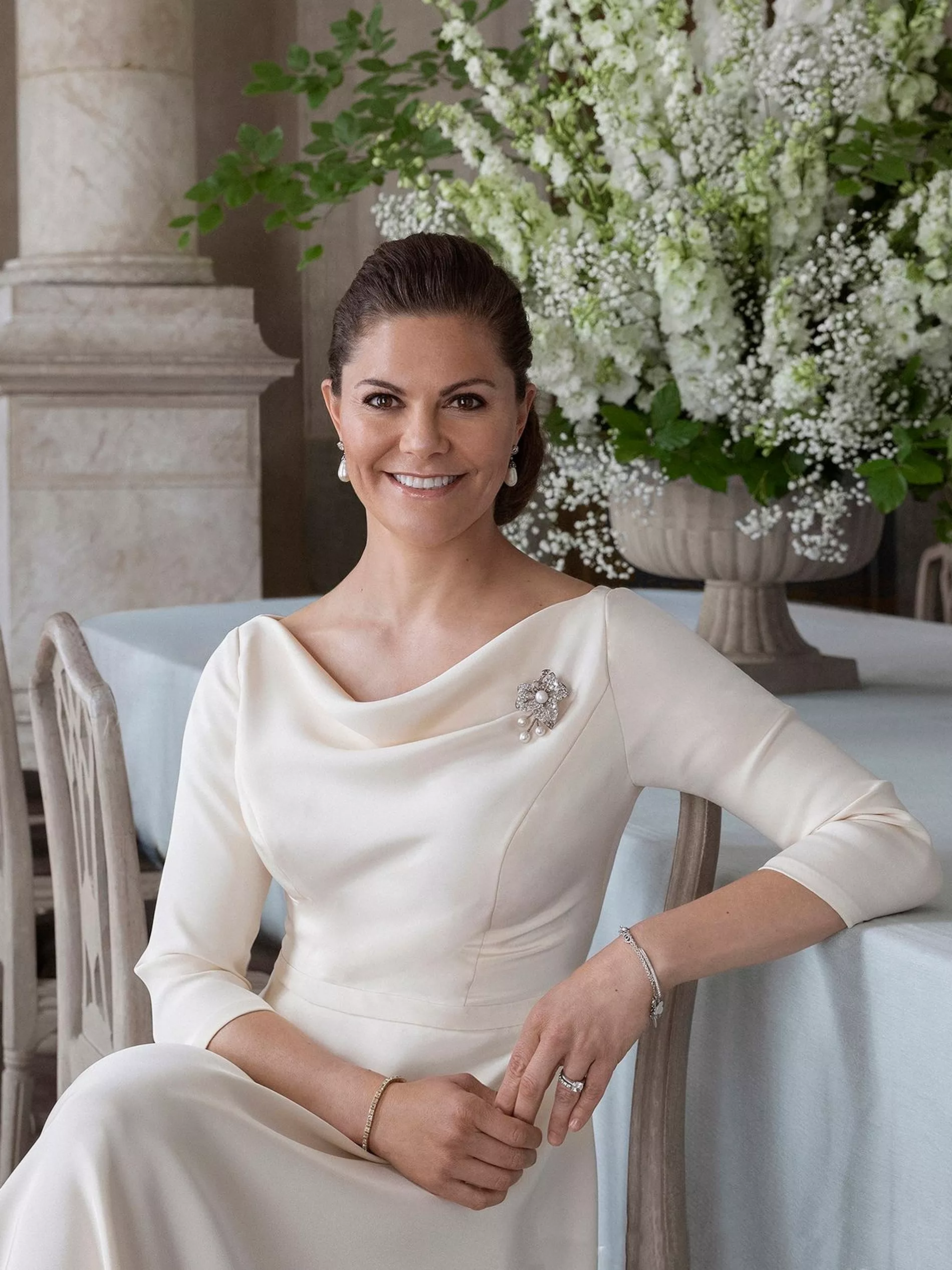 Crown Princess Victoria of Sweden poses sitting in a white gown with a draped neckline in a castle environment 