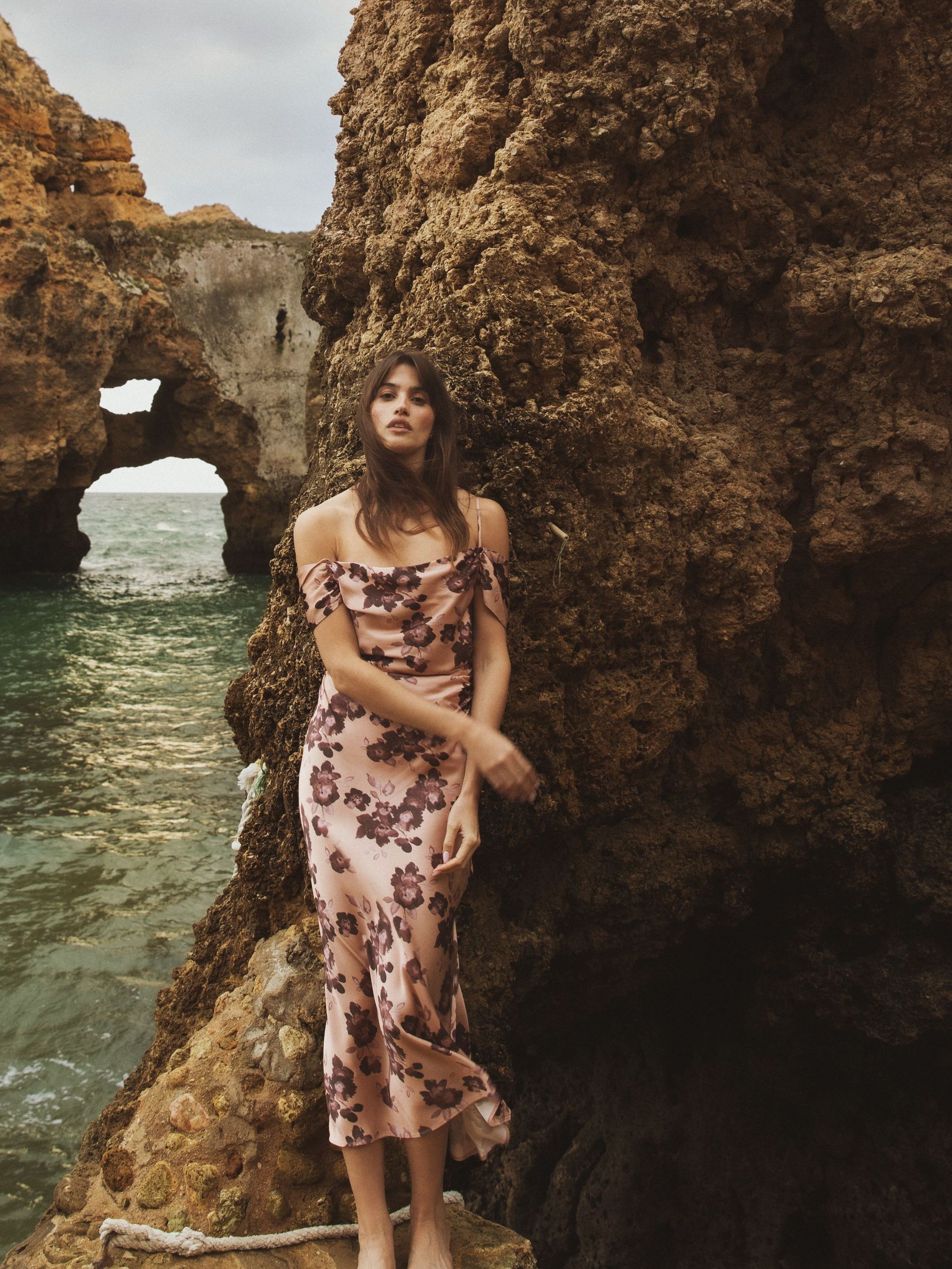 A women leaning against a cliff wall by the water, wearing a silk floral dress in dusty pink with dark flowers 