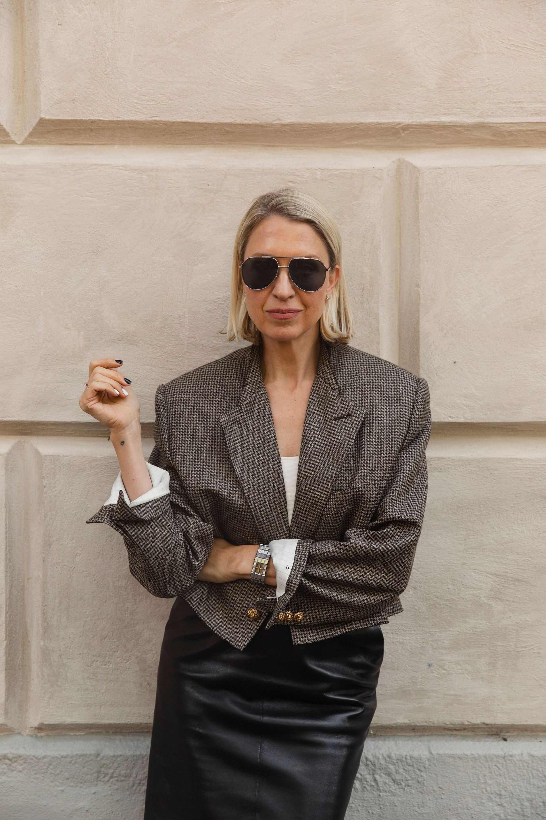 Emilia de Poret poses against a wall, wearing a houndstooth blazer, a white top, aviator sunglasses and a leather pencil skit