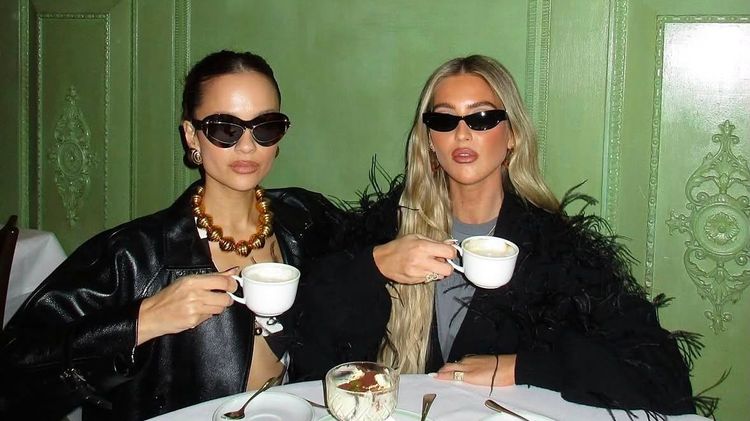 Emili Sindlev with friend drinking coffee wearing both black sunglasses and black jackets, the café has yellow and green walls with cornice, the table has a white cloth and dishes