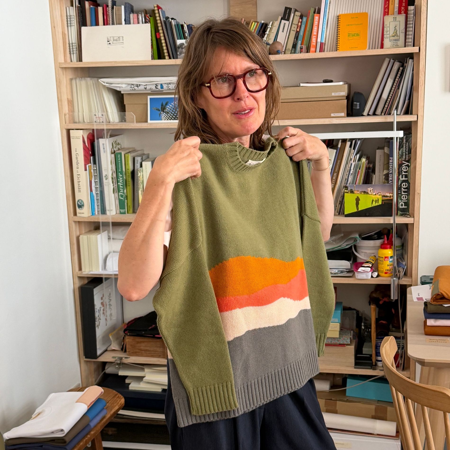 Caroline Hélain holding up the landscape-patterned sweater from her Longchamp collaboration in her Brussels studio, in front of shelves of sketchbooks and reference books.