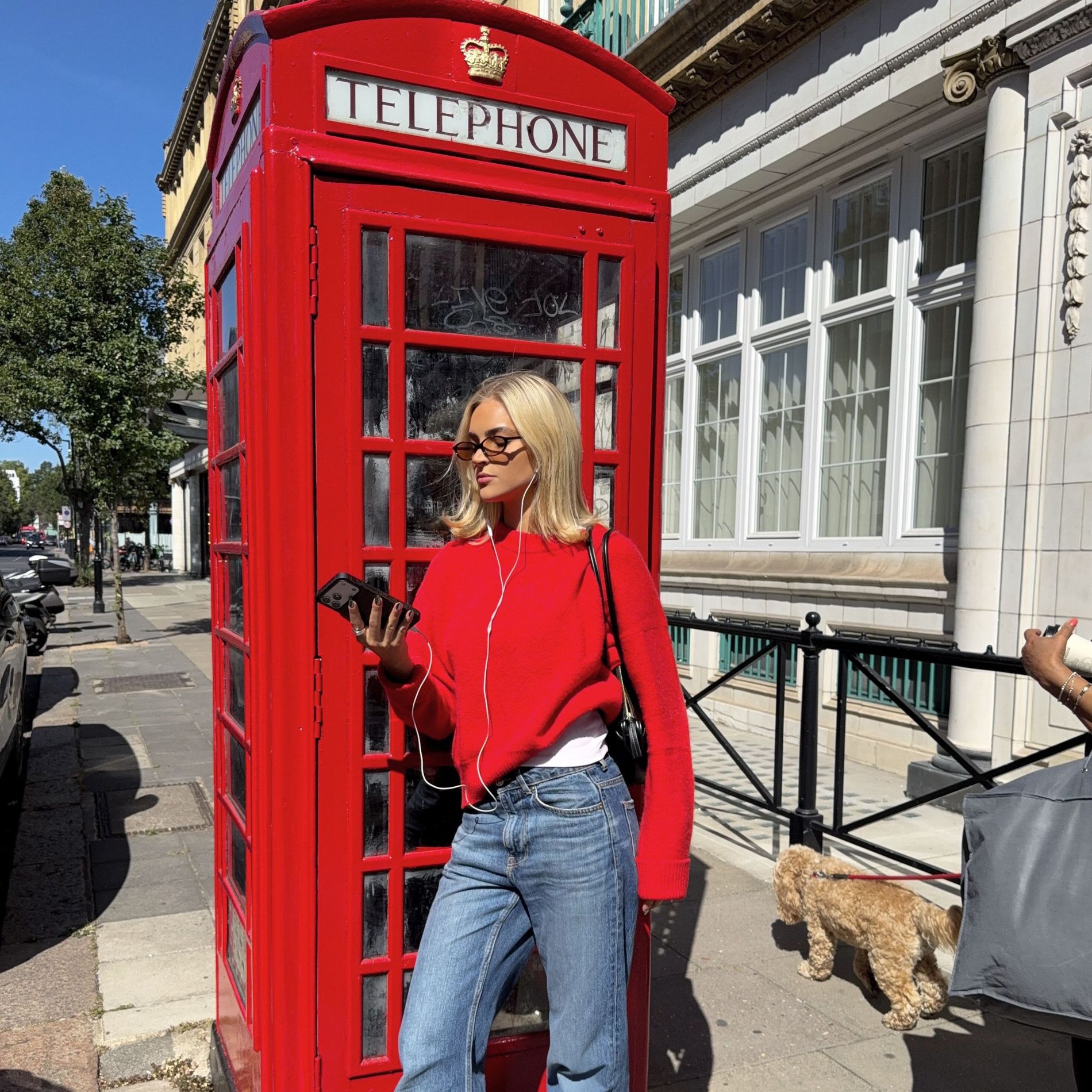 Emilia wears balck flipflops, jeans, and a red jumper by a London telephone box