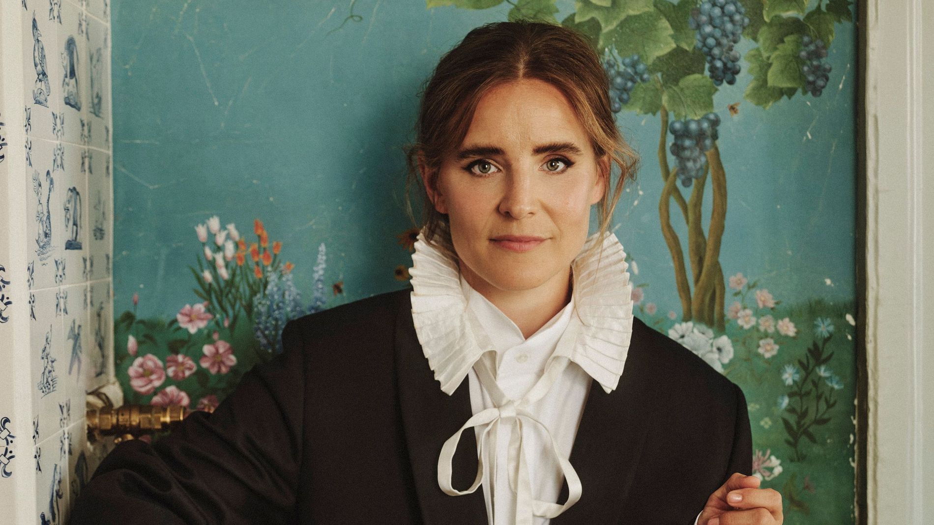 Beate Heuman stands against a teal wall with grape vine motif. She wears a black blazer, white shirt, and attached collar