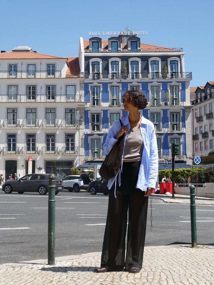 Swedish Malaika Holmen wears a grey tank top, a blue shirt, a suede bag and sheer black trousers posing on a street in Lisbon, Portugal