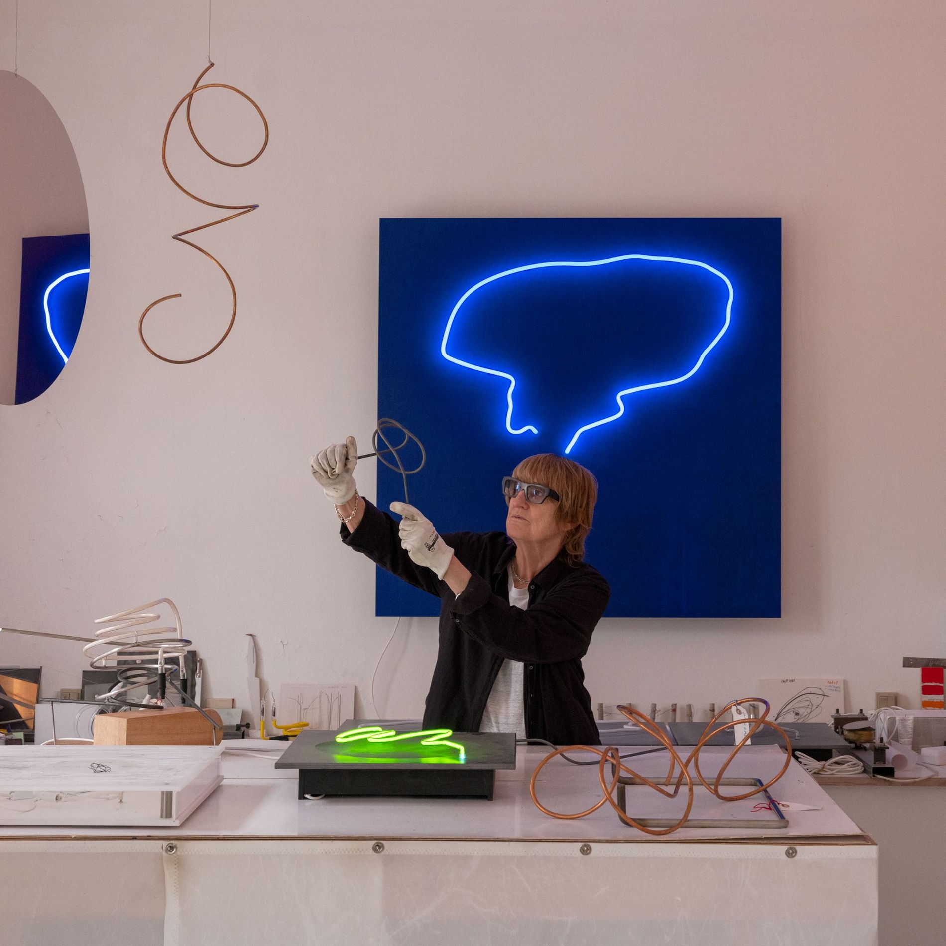 Gun Gordillo standing behind a desk with exam glasses working on a new creation, behind her is a dark blue painting and a neon blue loop shines