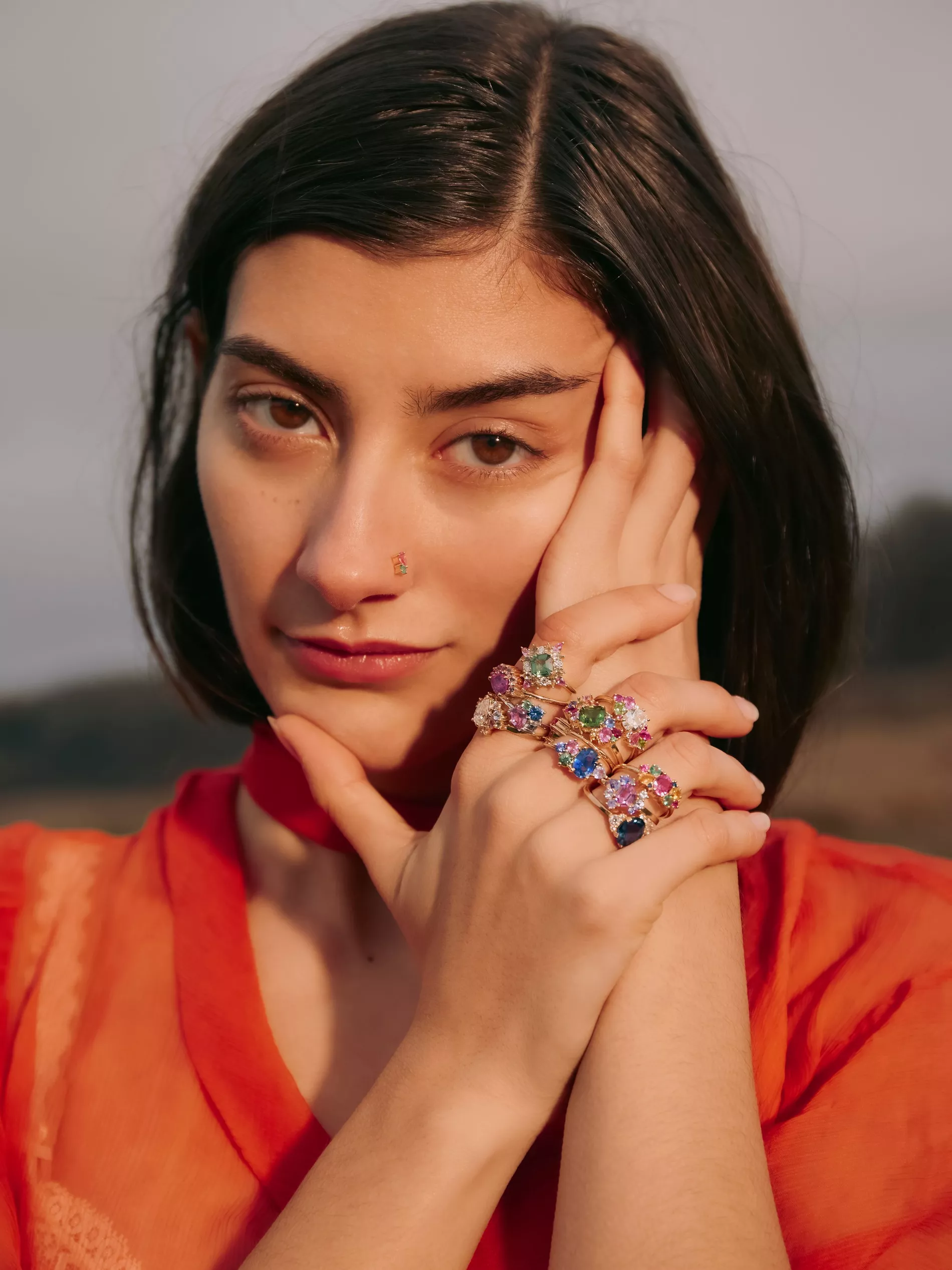 Up-close model posing with resting hands decorated with colourful gemstones from Anpé Atelier she is wearing a red-orange loose fitting top
