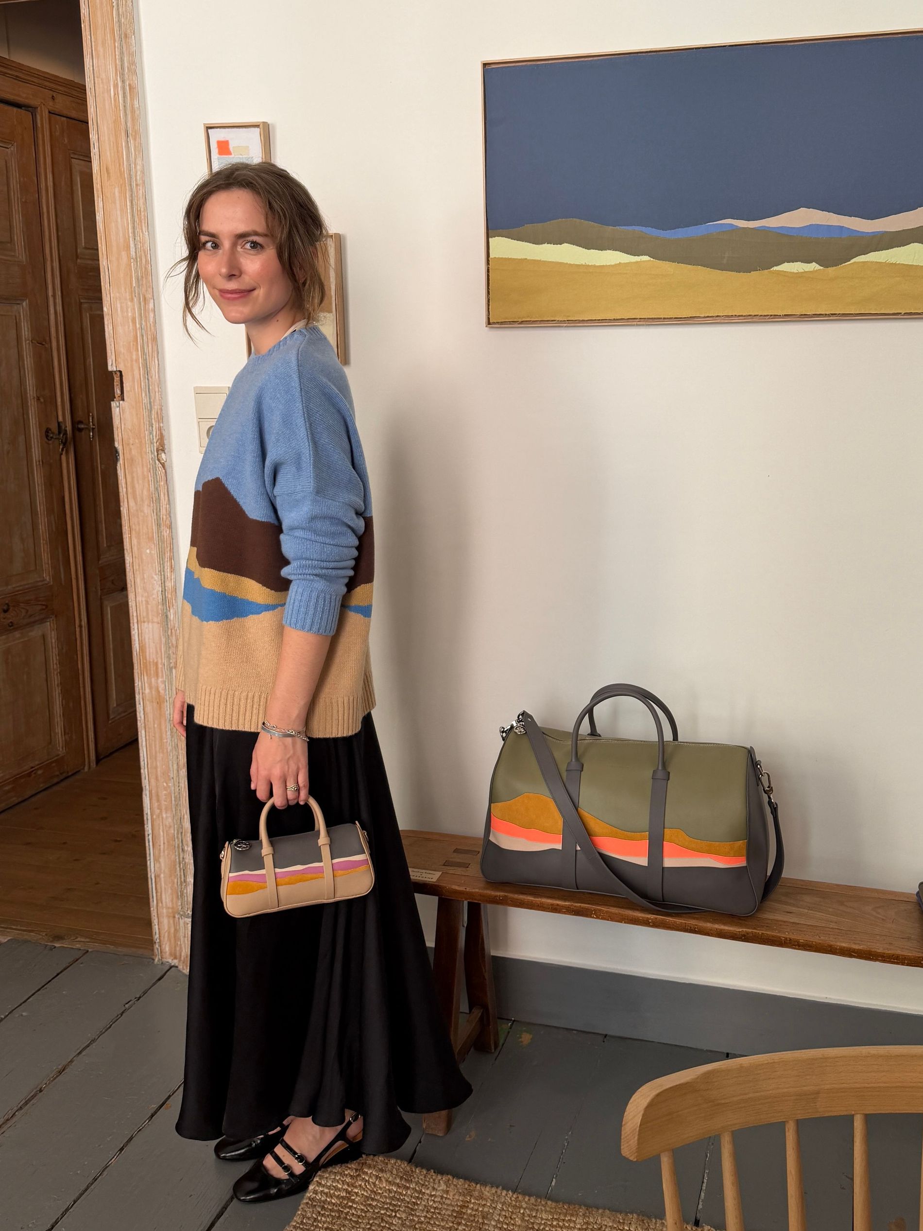 The writer wearing the landscape-print sweater and taupe Daylong mini bag from the Caroline Hélain x Longchamp collection, with the olive green Daylong weekender on the bench behind her.