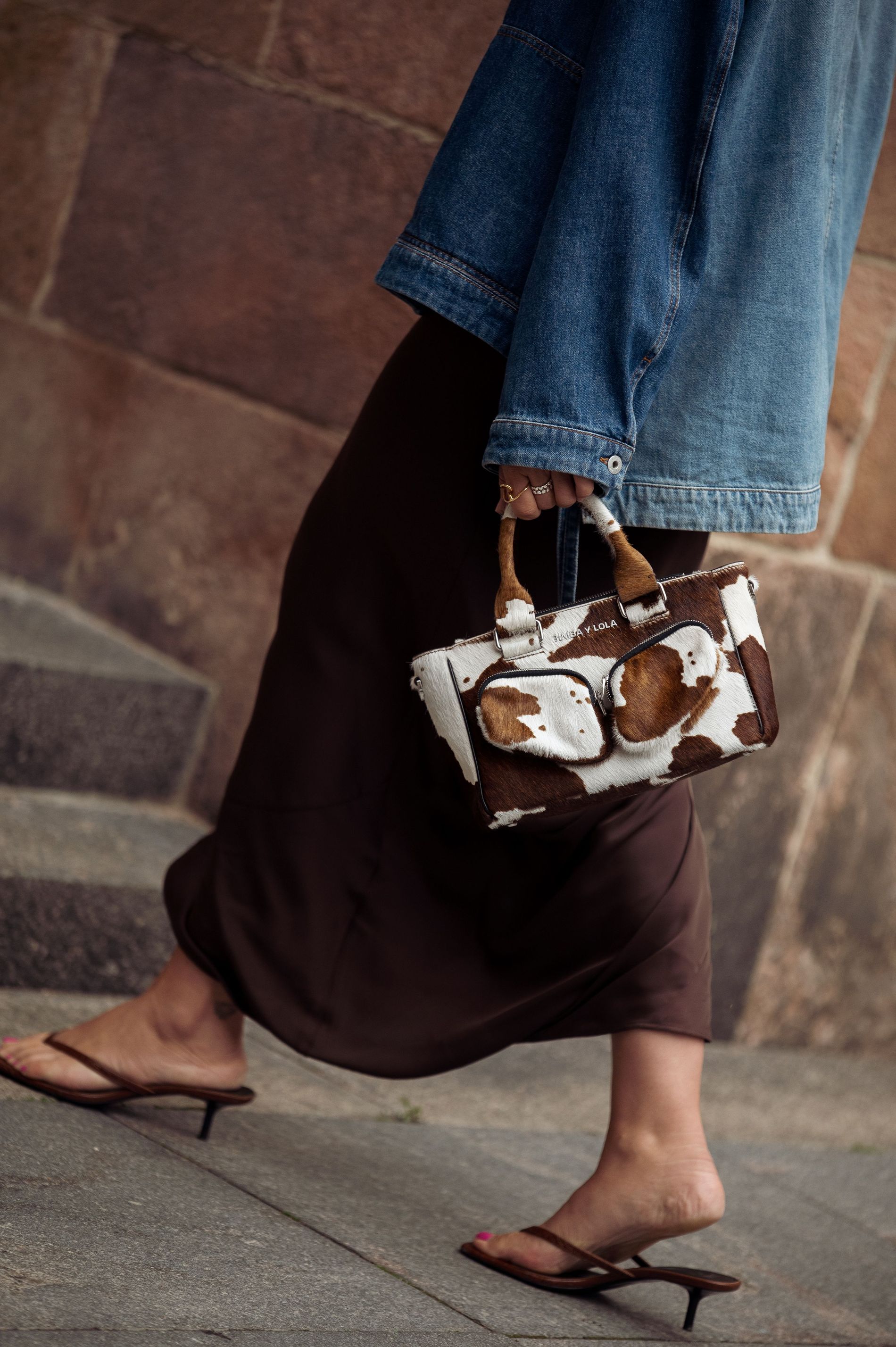 Close-up of a brown and white cow print mini bag styled with a brown slip skirt, kitten heels, and a denim jacket