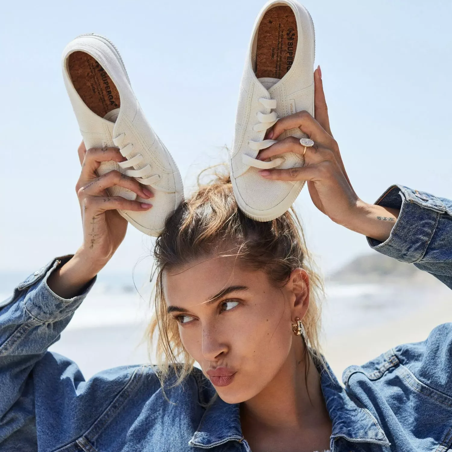 Hailey Bieber standing on a beach wearing a jean jacket and white top with white sneakers on top of her head