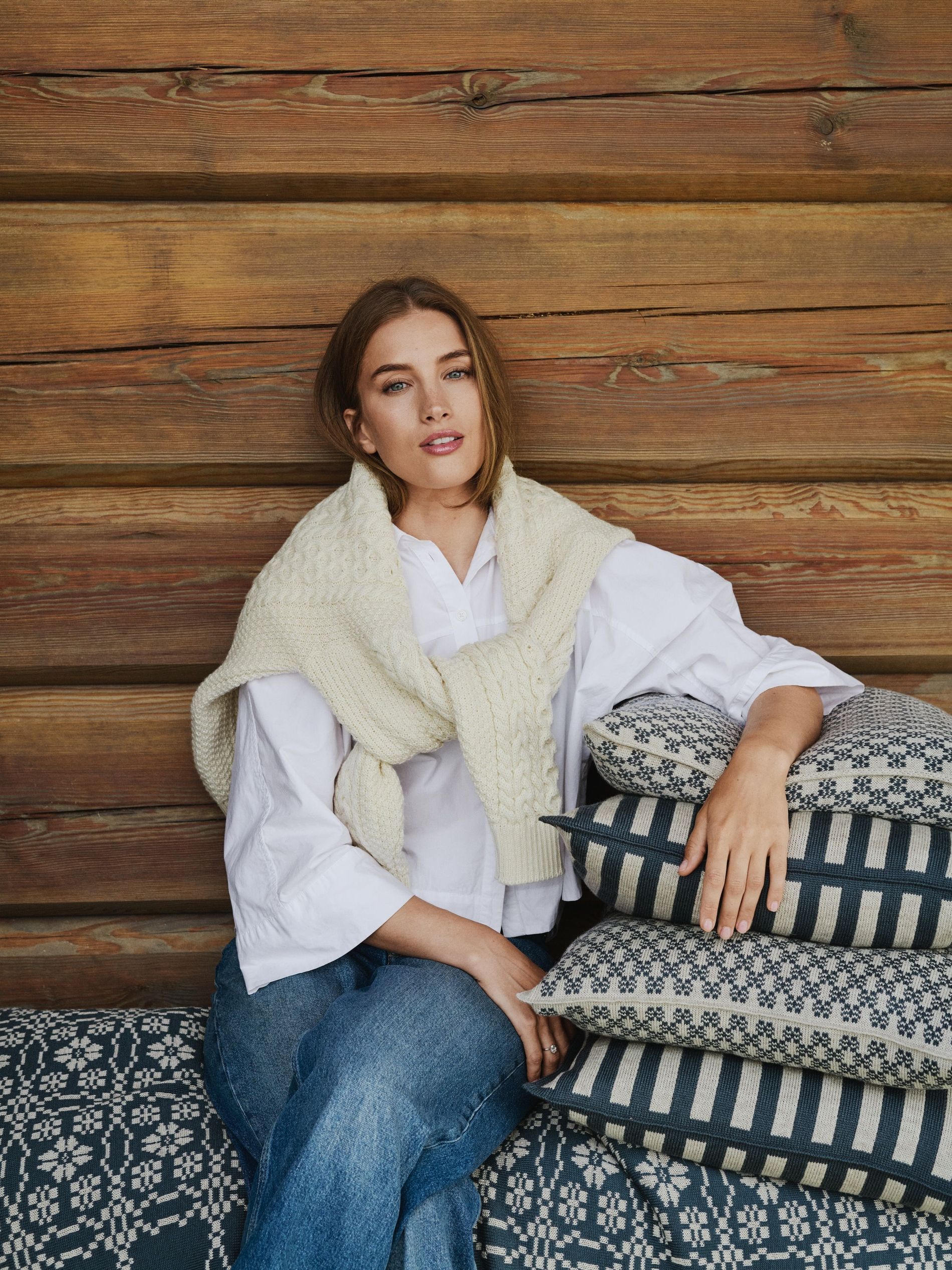Pernille Døsvik sitting with her piles of Dales Of Norway pillows wearing blue jeans and a dale of norway white sweather and a white shirt