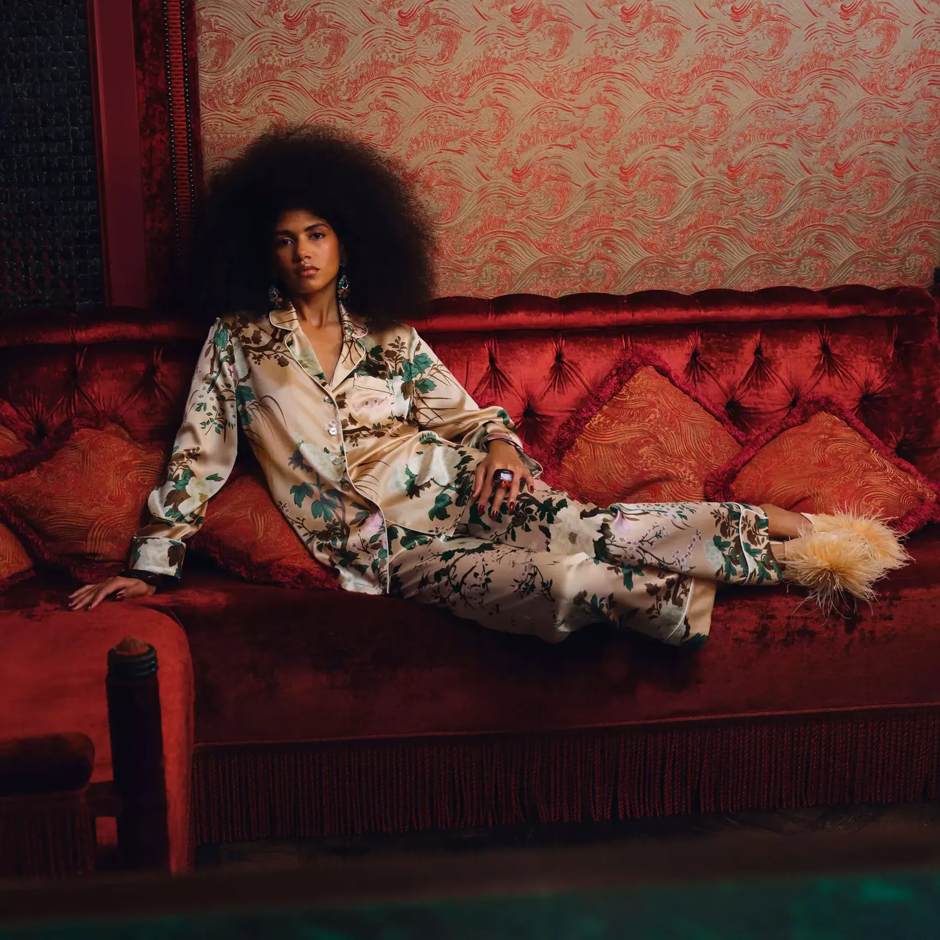 Woman leaning on a red velvet sofa wearing a patterned silk pyjama in pink with light pink feather shoes 