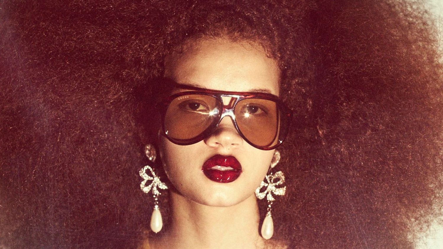 Model wearing brown shield glasses and sparkling drop earrings look straight at the camera