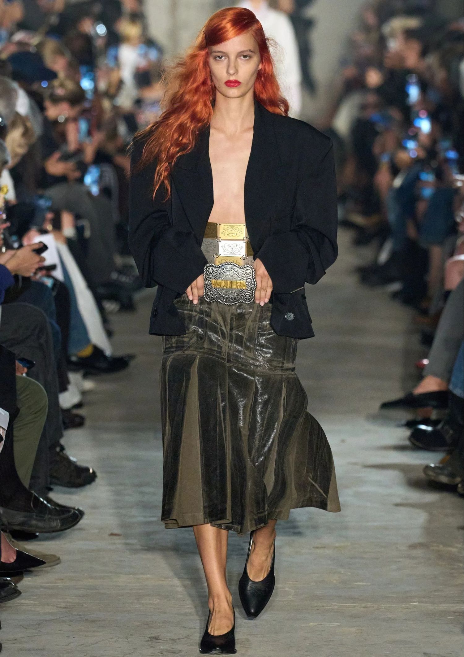 Model walking the Vaquera SS25 runway wearing midi skirt in green and black blazer, with stacked belts in gold and silver 