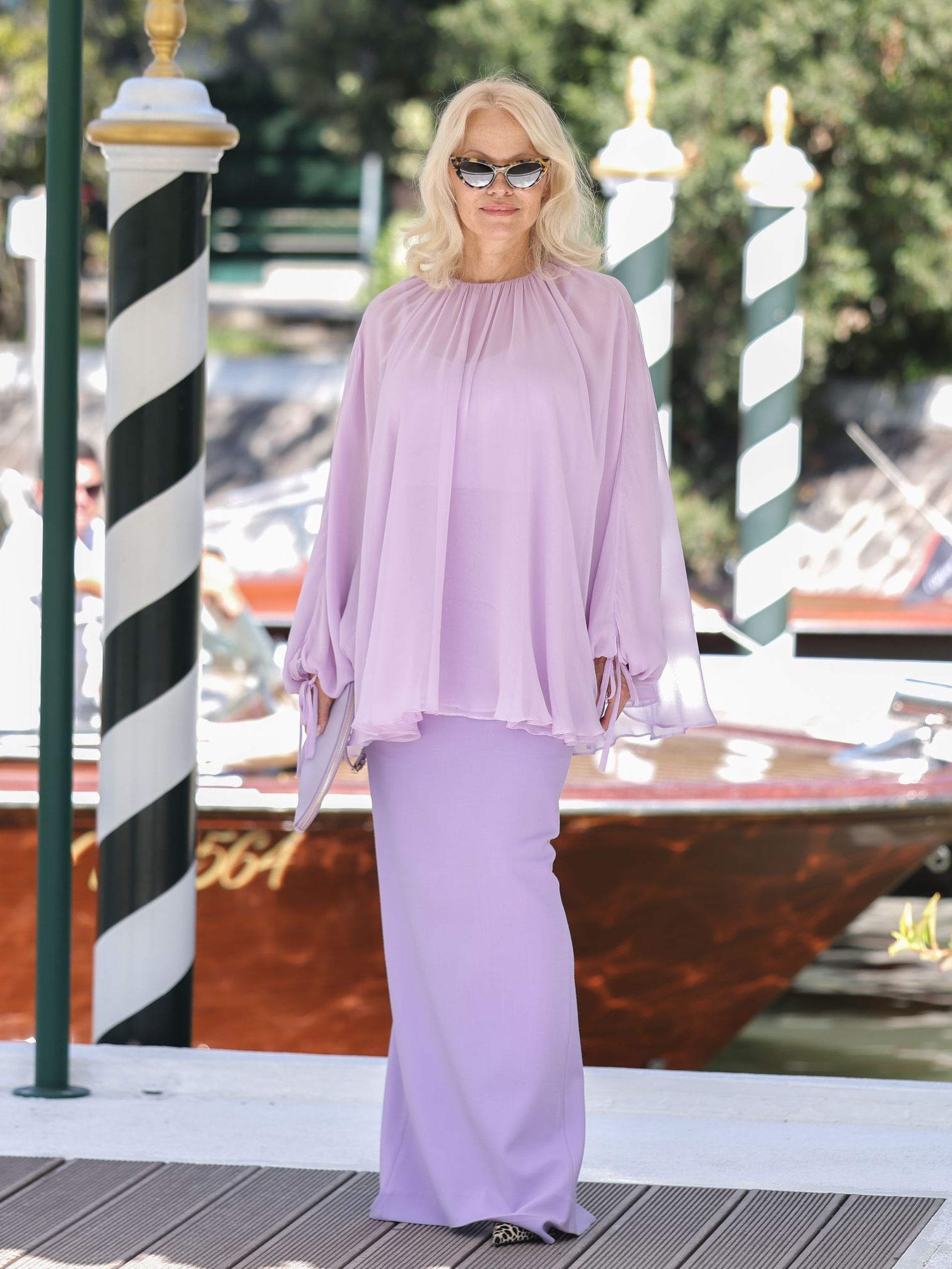 Pamela Anderson stands on the dock at the Venice Film Festival wearing a lilac two piece and black sunglasses