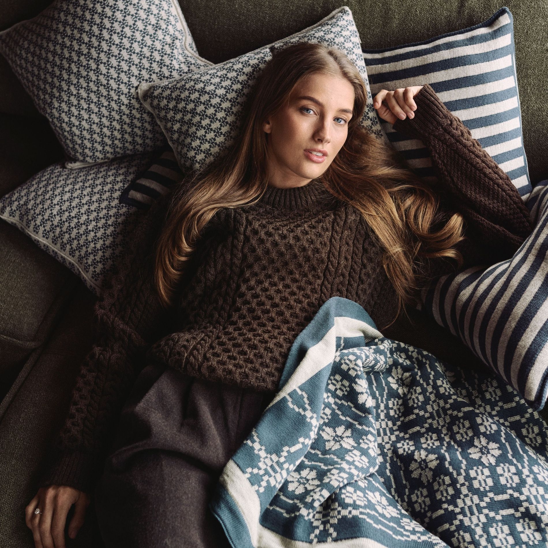 Pernille Dosvik lounges in a Dale of Norway sweater on the sofa with pillows and a blanket from her Dale of Norway collaboration