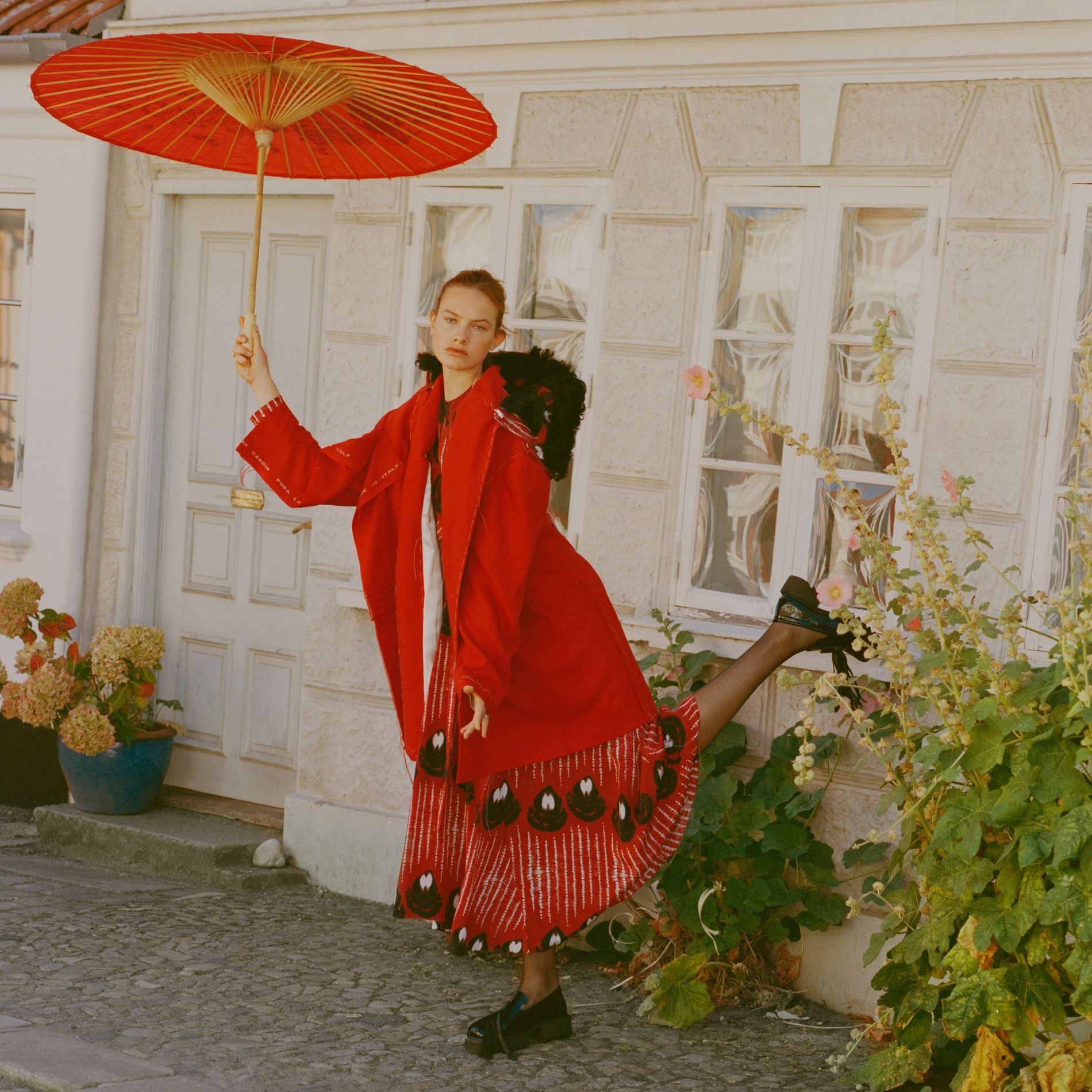 Model stands in a courtyard in a red coat, red skirt and black shoes holding a red umbrella