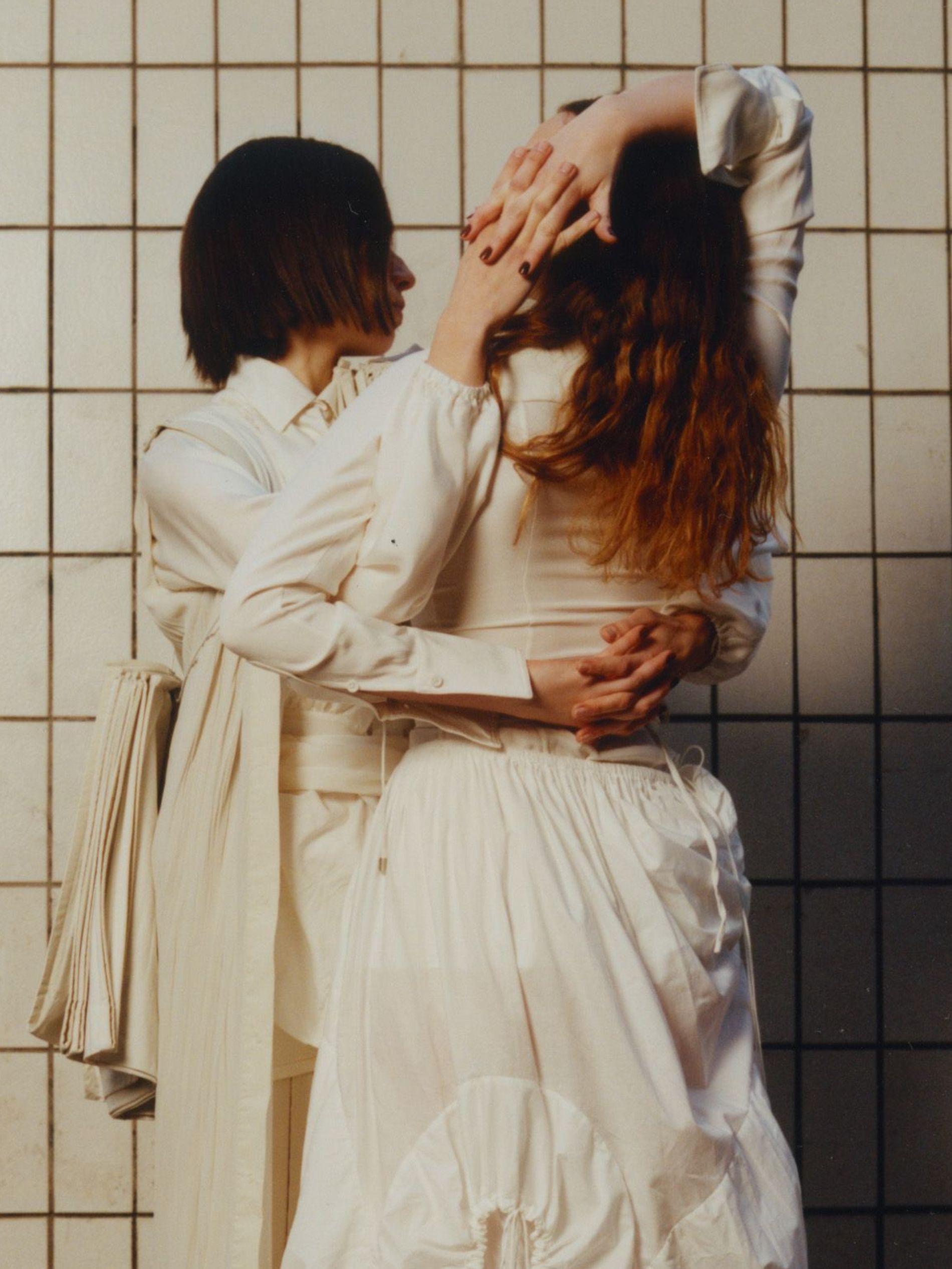 Two women holding hands behind their backs in a tiled room wearing all white