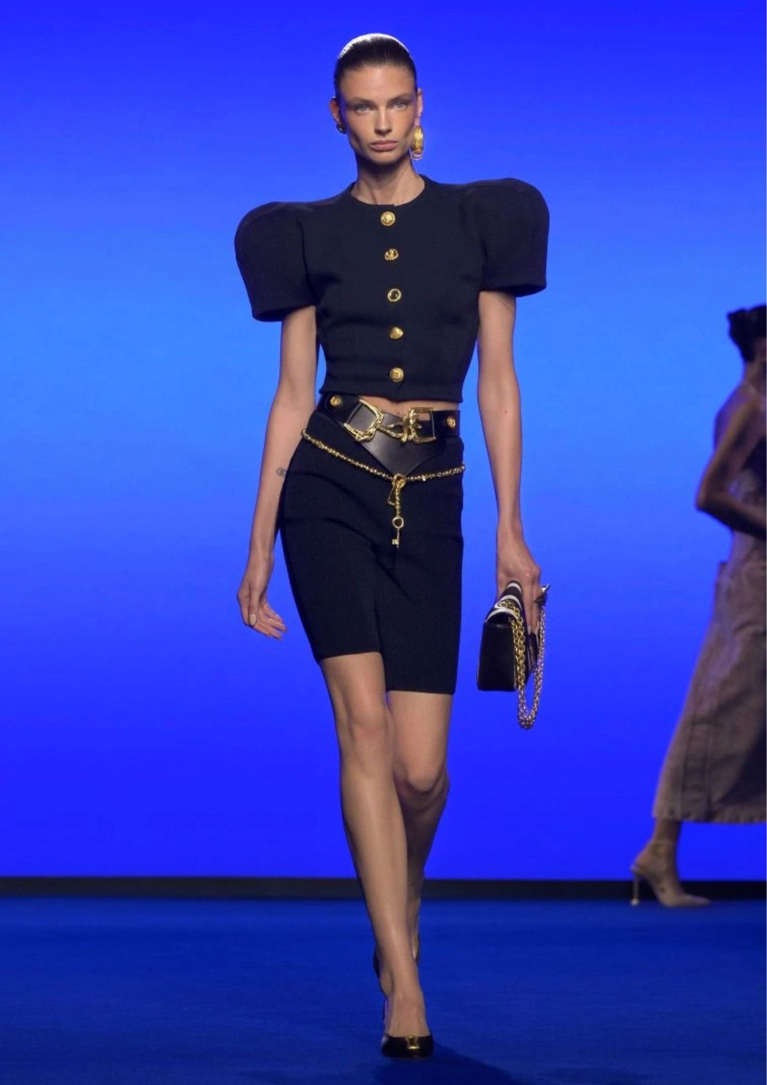 Model walking the Schiaparelli SS25 runway, wearing black ensemble with stacked belts in black leather and gold chain 