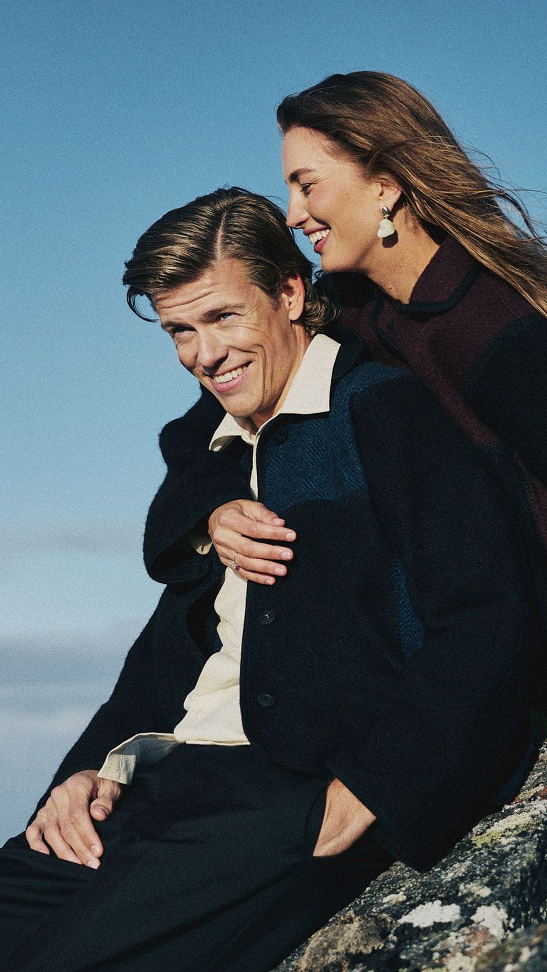 Klæbo and Døsvik sit on a rock. He wears black trousers and jacket with denim details with cream shirt. She wears all black