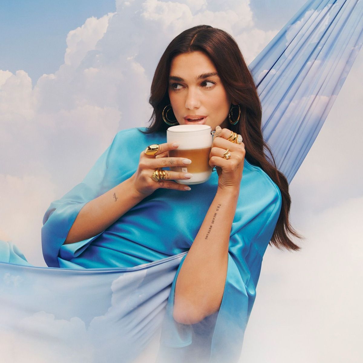 Dua Lipa announced as Nespresso's brand ambassador. The image sees her suspended in the clouds, in a hammock, a cup of frothy coffee in her hand. She is dressed in a blue satin gown from Pinko