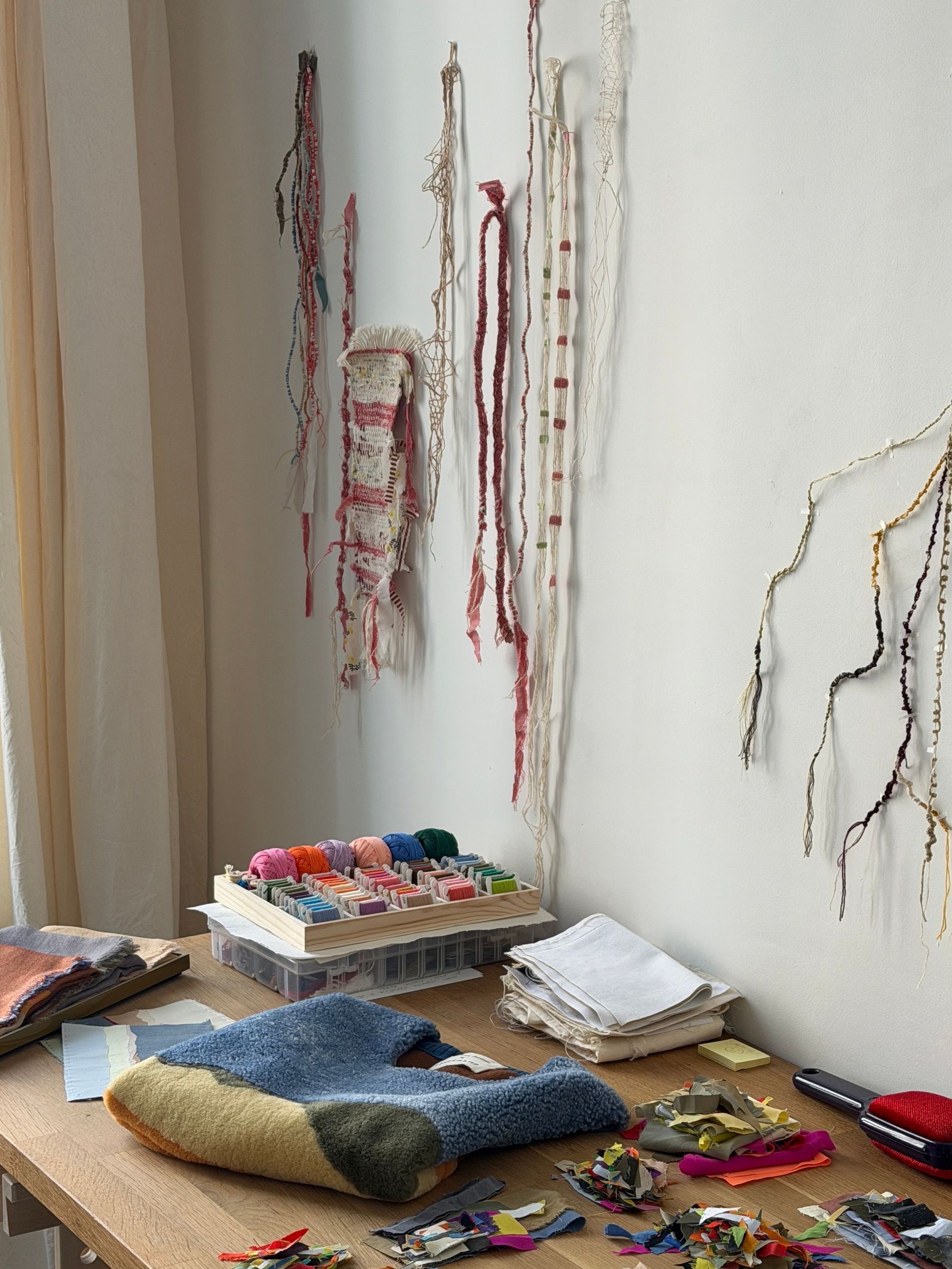 Experimental thread and fabric studies hanging on the wall of Caroline Hélain's Brussels studio, above a desk holding the sleeveless top from her Longchamp collaboration and fabric scraps sorted by colour.