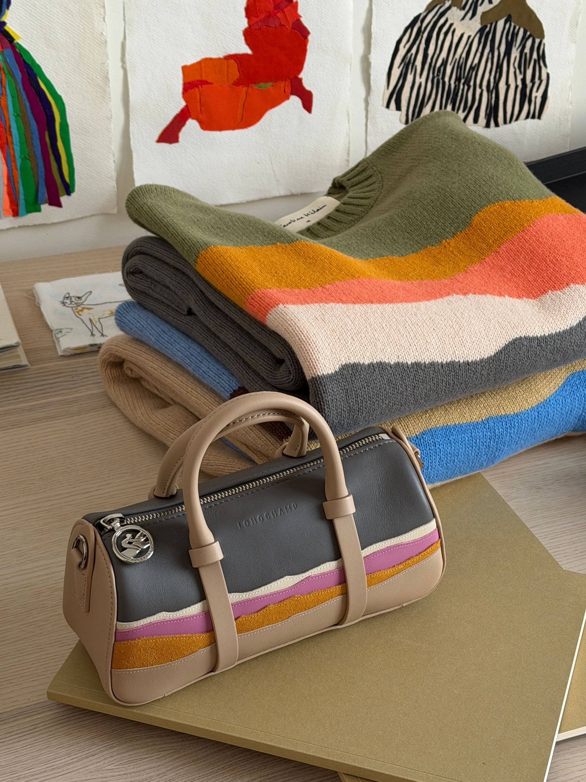 Caroline Hélain's Brussels studio desk, with the Longchamp Daylong mini bag and folded knitwear from her collaboration displayed beside her original patchwork landscape artworks.