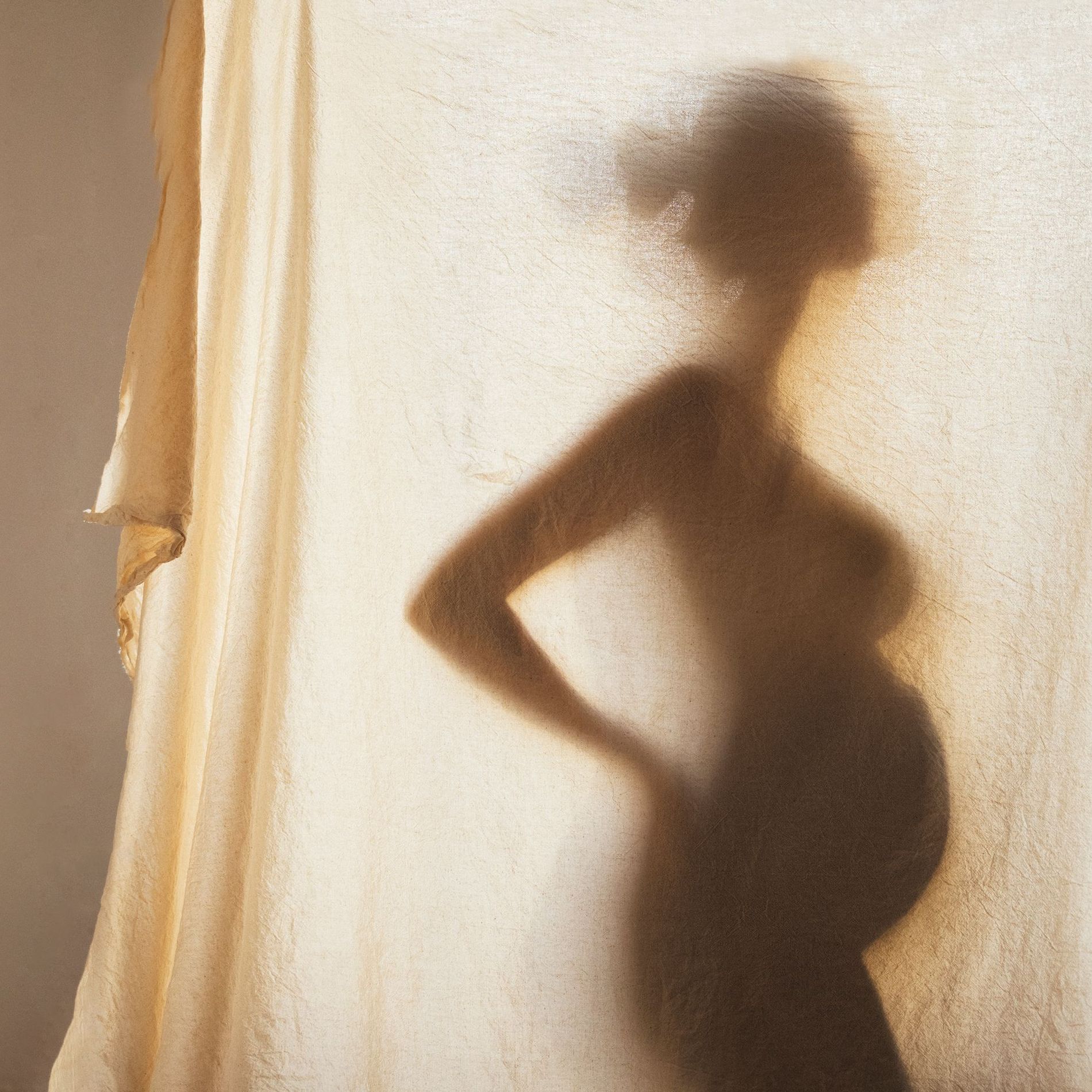 Silhouette of a pregnant woman is seen through a linen curtain