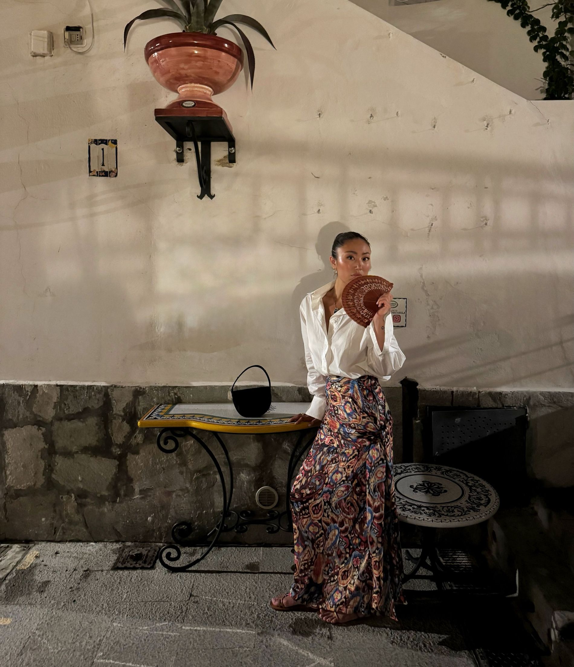 Alice Wang wears a patterned maxi skirt and white oversized shirt , with slicked back hair and an ornate fan