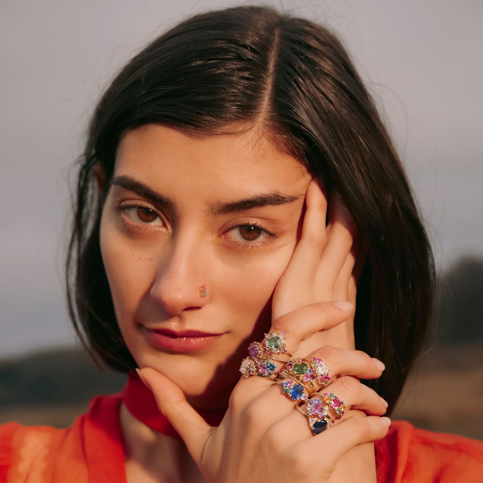 Up-close model posing with resting hands decorated with colourful gemstones from Anpé Atelier she is wearing a red-orange loose fitting top