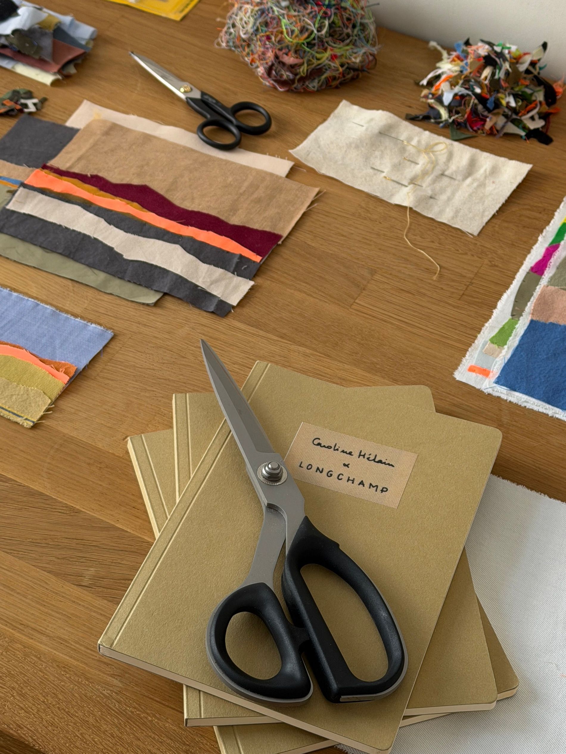 Caroline Hélain's fabric scissors resting on sketchbooks labelled "Caroline Hélain x Longchamp," surrounded by thread offcuts and hand-cut landscape patchwork swatches.