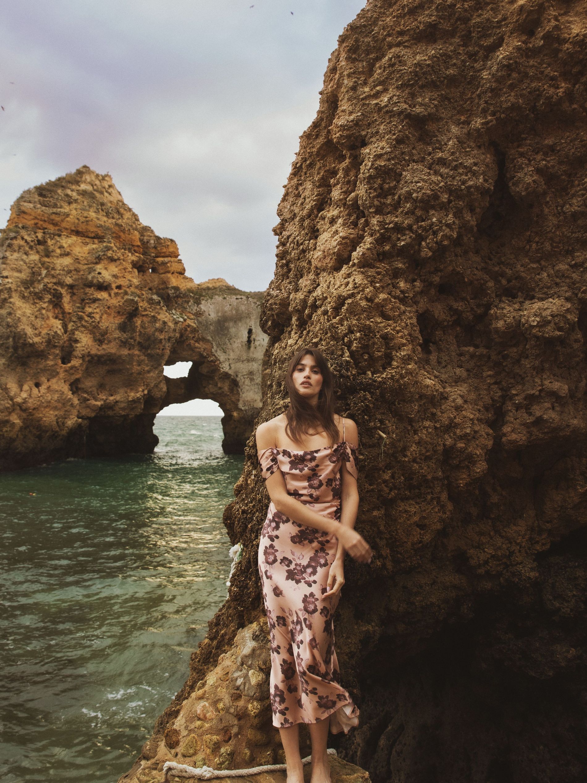 A women leaning against a cliff wall by the water, wearing a silk floral dress in dusty pink with dark flowers 