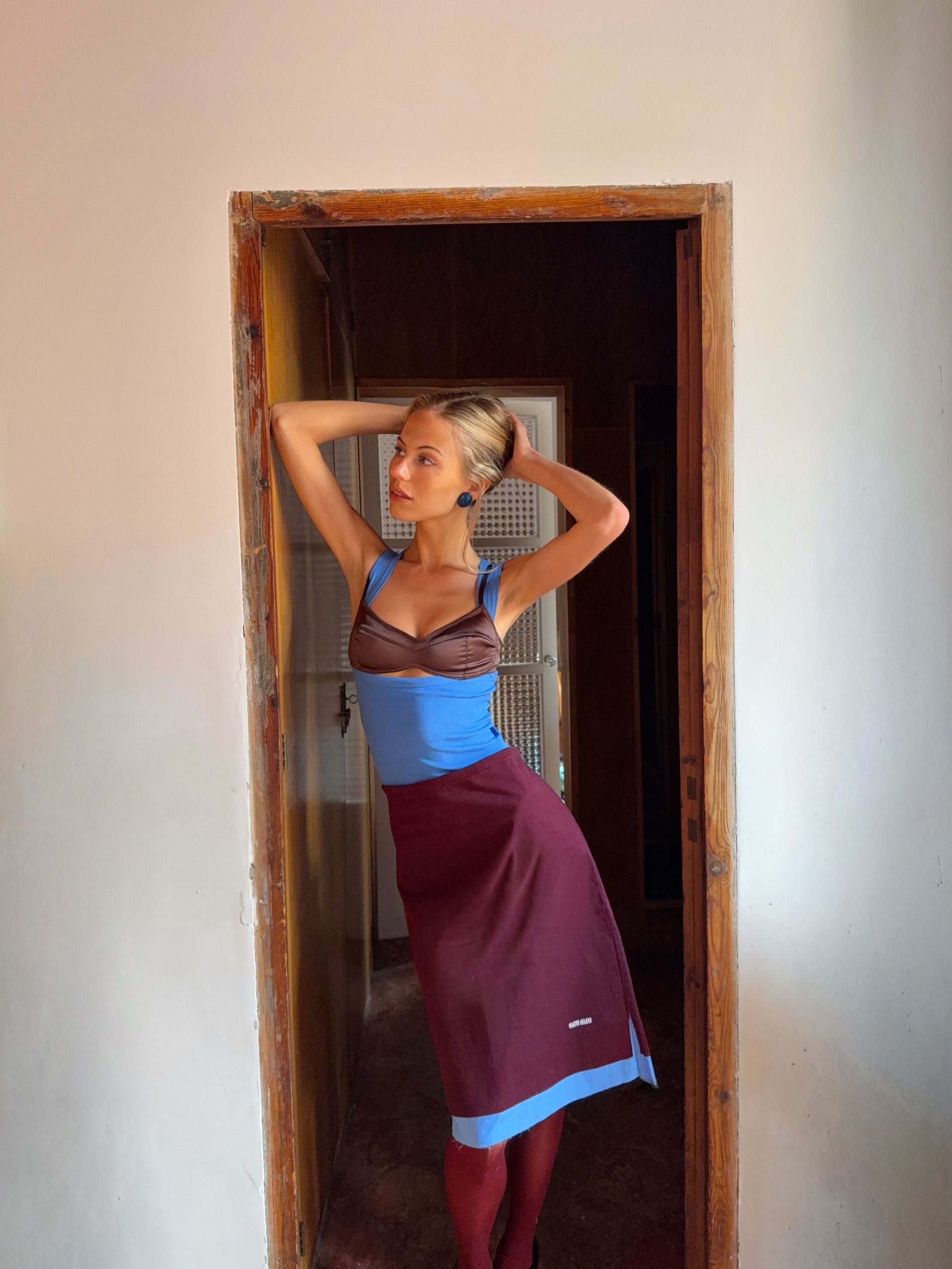 Influencer Elin Warn poses in a doorway on vacation wearing a brown and blue Gem Wear top and bra, a burgundy Miu Miu skirt with matching Swedish Stockings tights, Prada shoes and her grandma's blue earrings