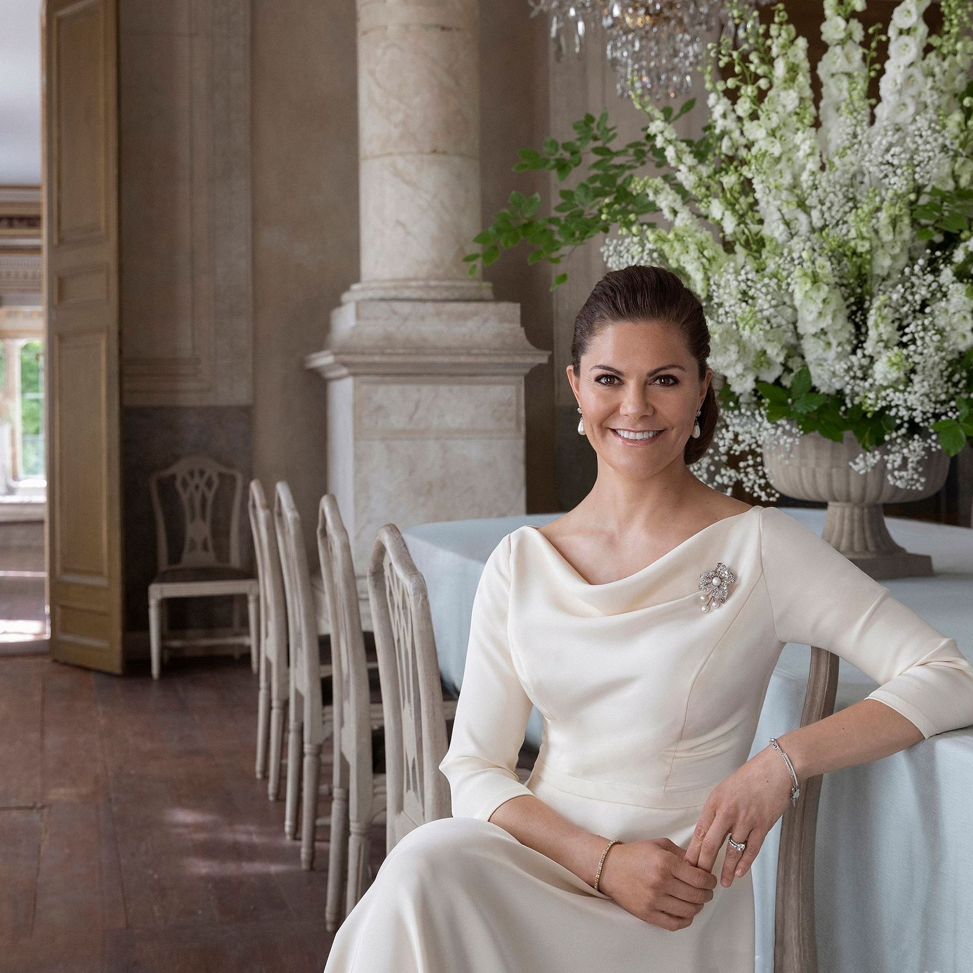 Crown Princess Victoria of Sweden poses sitting in a white gown with a draped neckline in a castle environment 