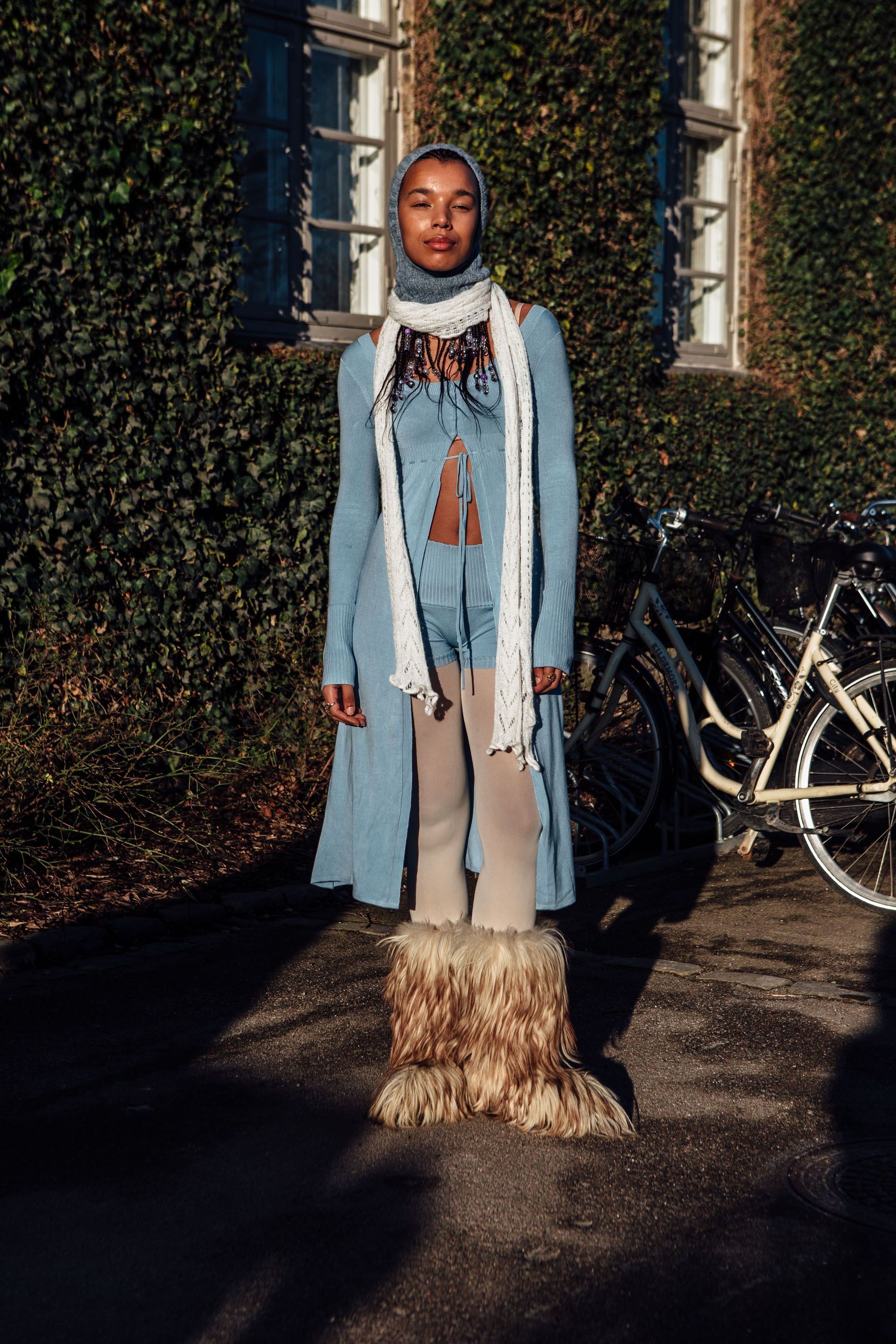 A street style guest at CPHFW AW23 wears a grey balaclava, a white skinny scarf, a knitted set and faux fur boots