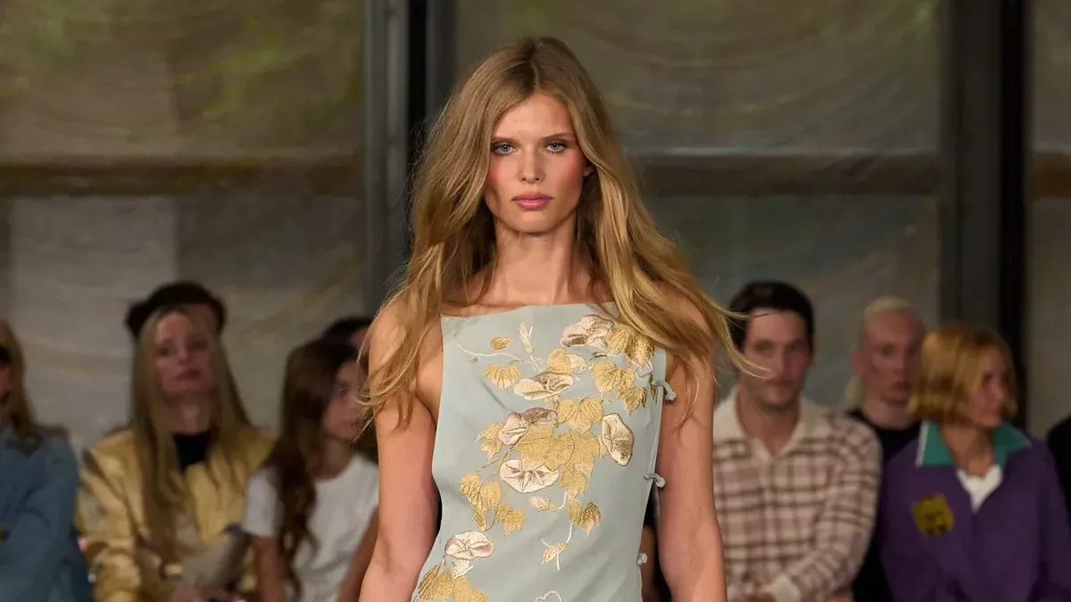Ida Heiner walks Conner Ives' SS27 show at New York Fashion Week. She wears a blue dress and neon yellow stoll, walking a carpeted runway.