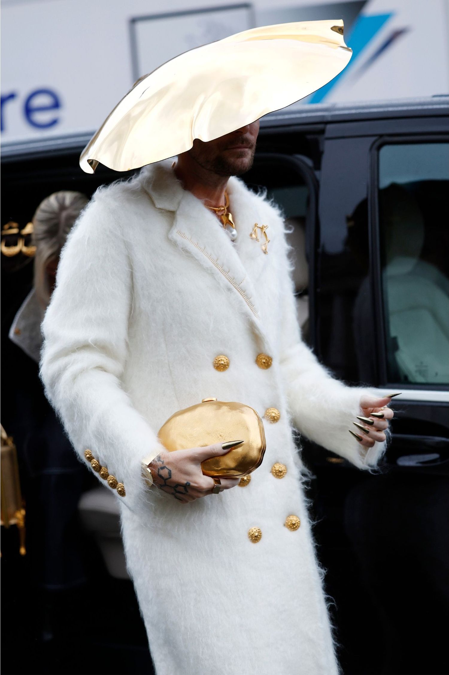 Fredrik Robertsson wears cream Schiaparelli coat, black metallic Schiaparelli bag, gold metallic large hat, outside Schiaparelli, during Haute Couture Spring-Summer 2025