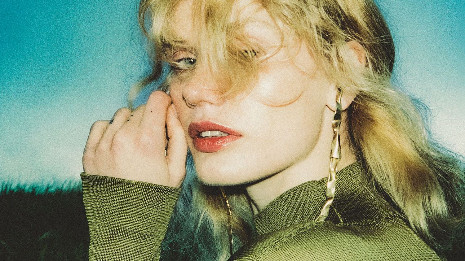 Model with tousled blonde hair and glowing skin looks at the camera framed by the setting summer sun