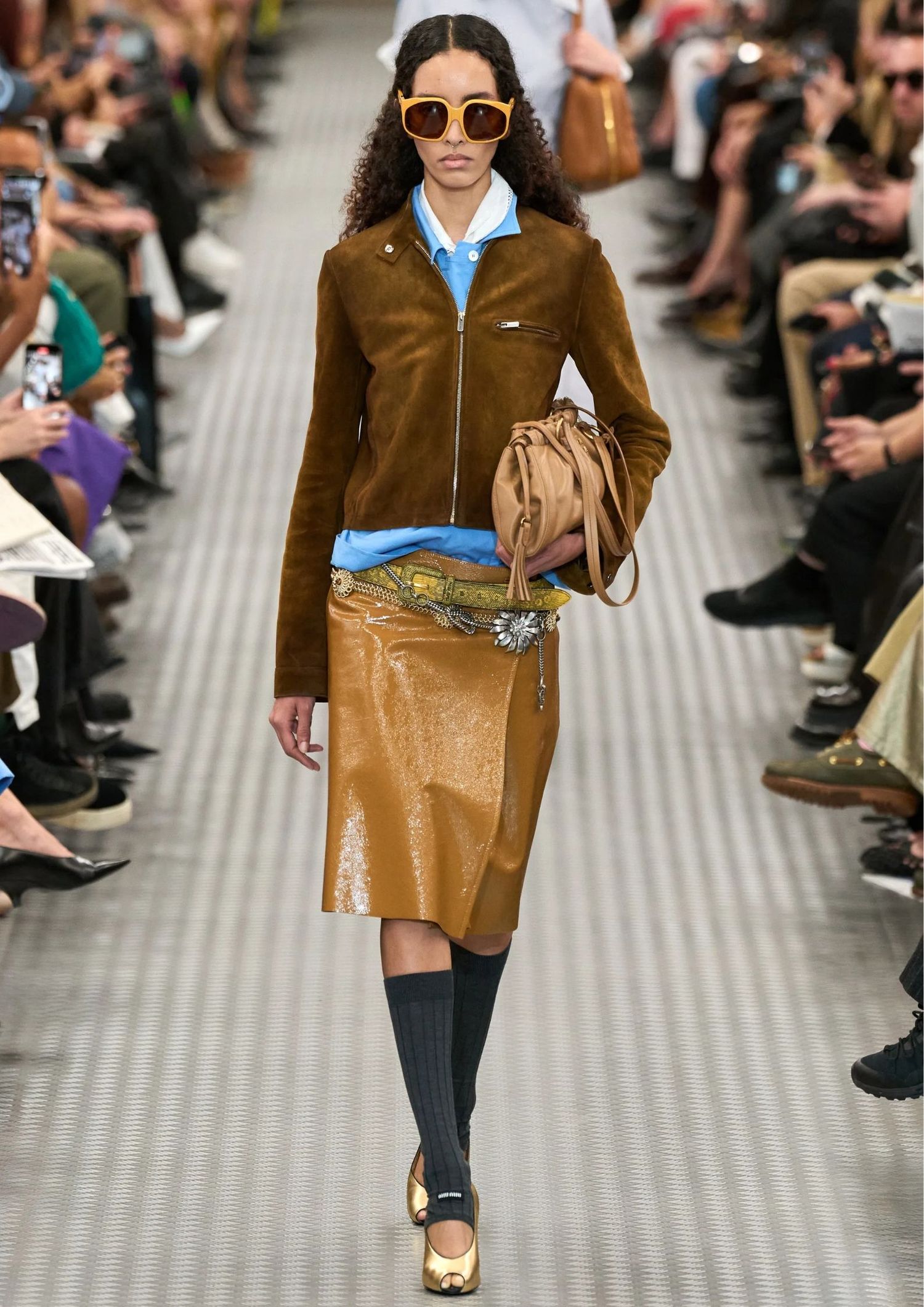 Model walking the Miu Miu SS25 runway, wearing patent leather skirt, brown suede jacket and stacked belts  