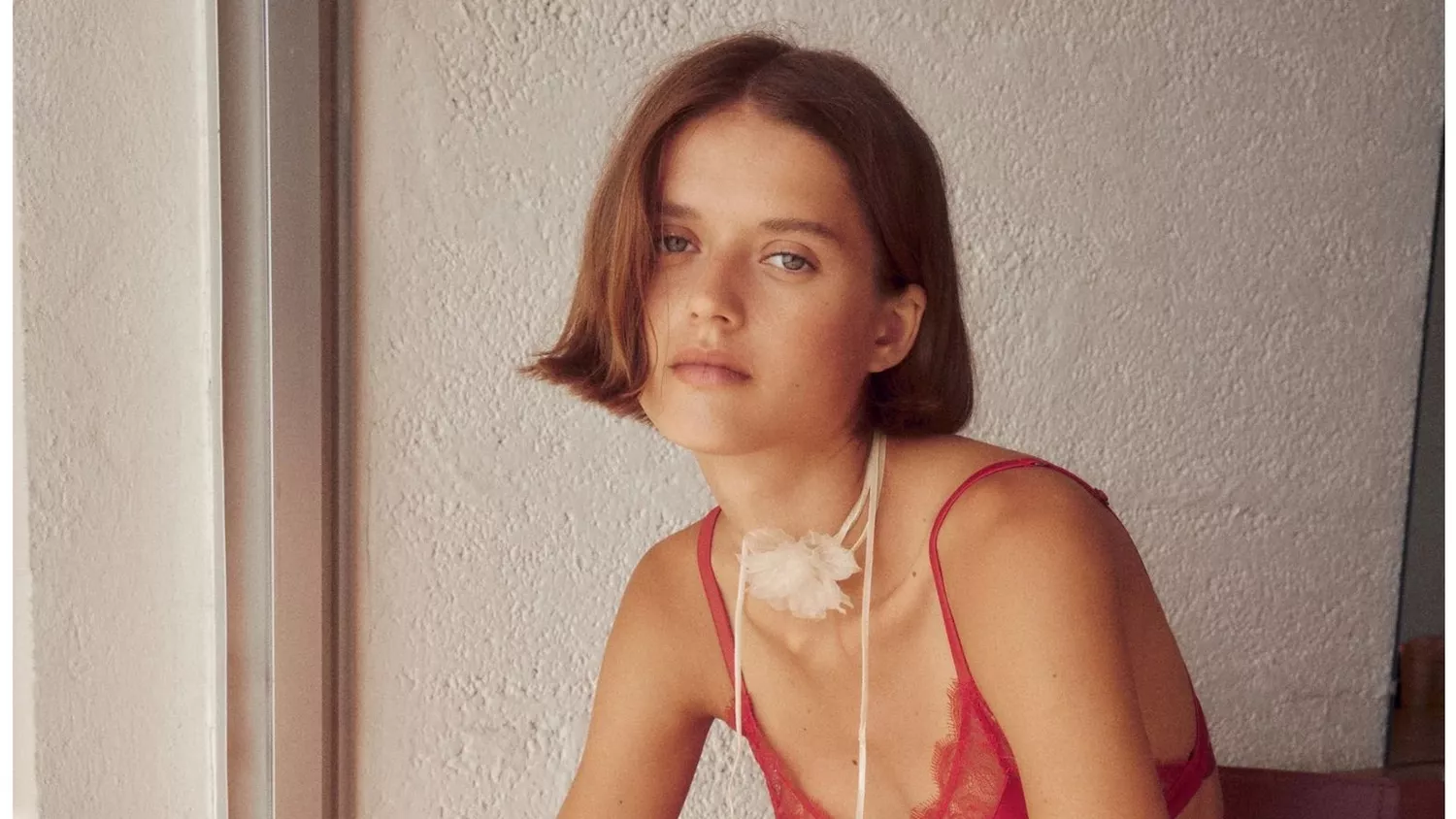 Woman sitting in chair wearing red lace bra, lace briefs and a white choker with flower 