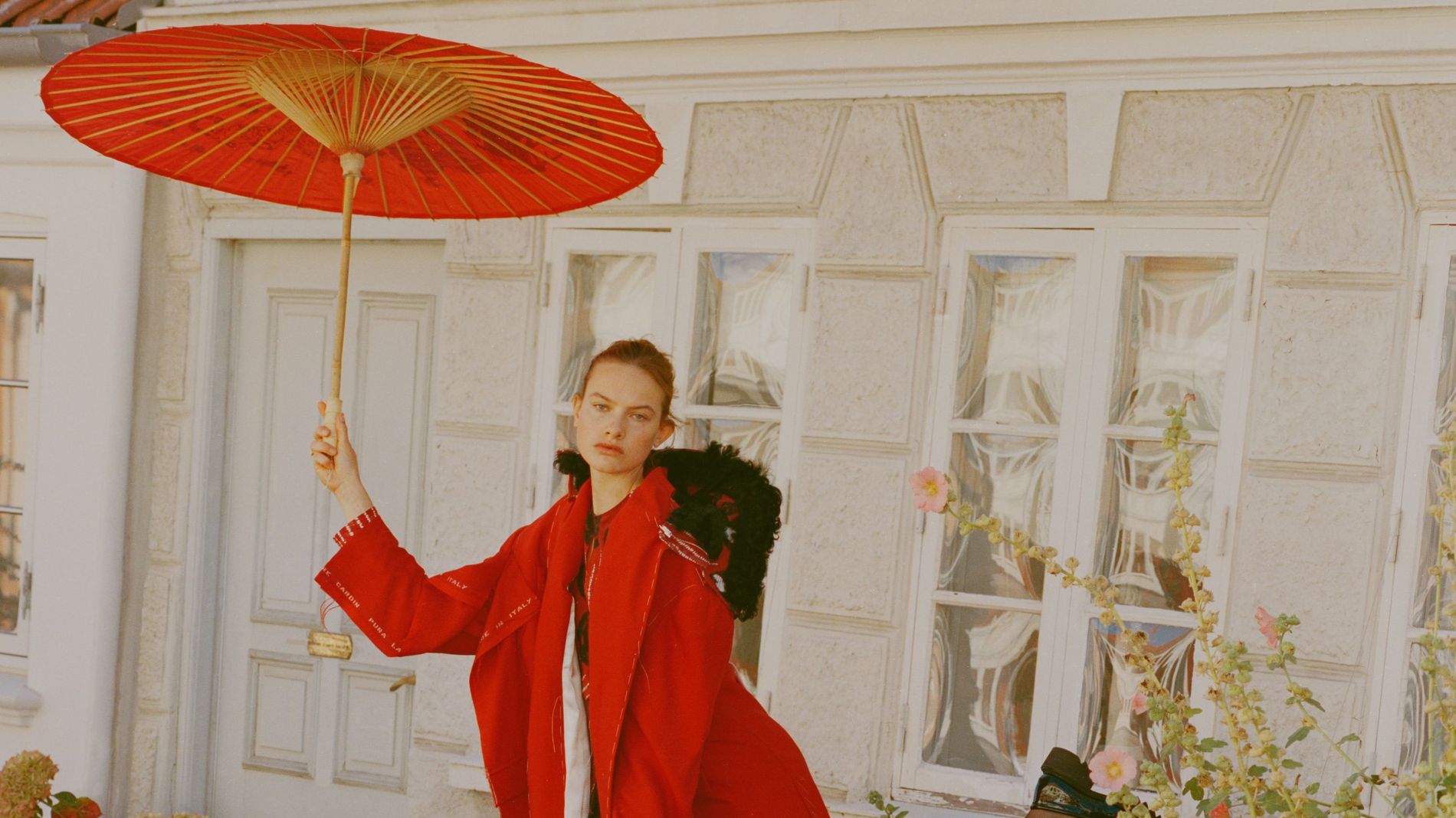 Model stands in a courtyard in a red coat, red skirt and black shoes holding a red umbrella