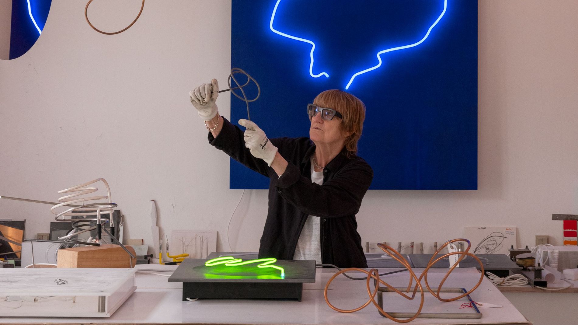 Gun Gordillo standing behind a desk with exam glasses working on a new creation, behind her is a dark blue painting and a neon blue loop shines