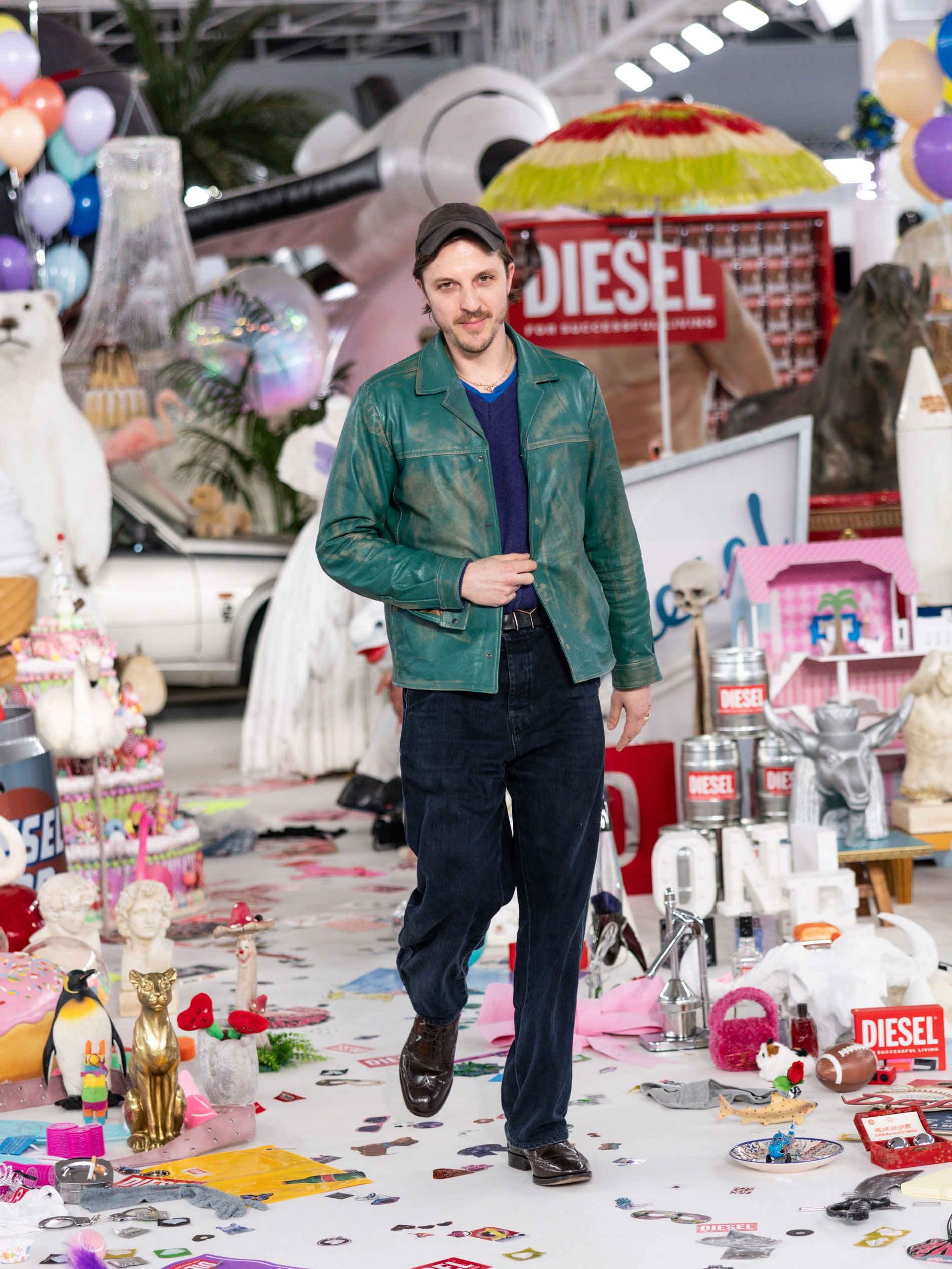 Glenn Martens wearing a green patrol leather jacket and dark blue jeans with brown shoes matching with a brown cap, he has alot of junk behind him from one of Diesels runway shows